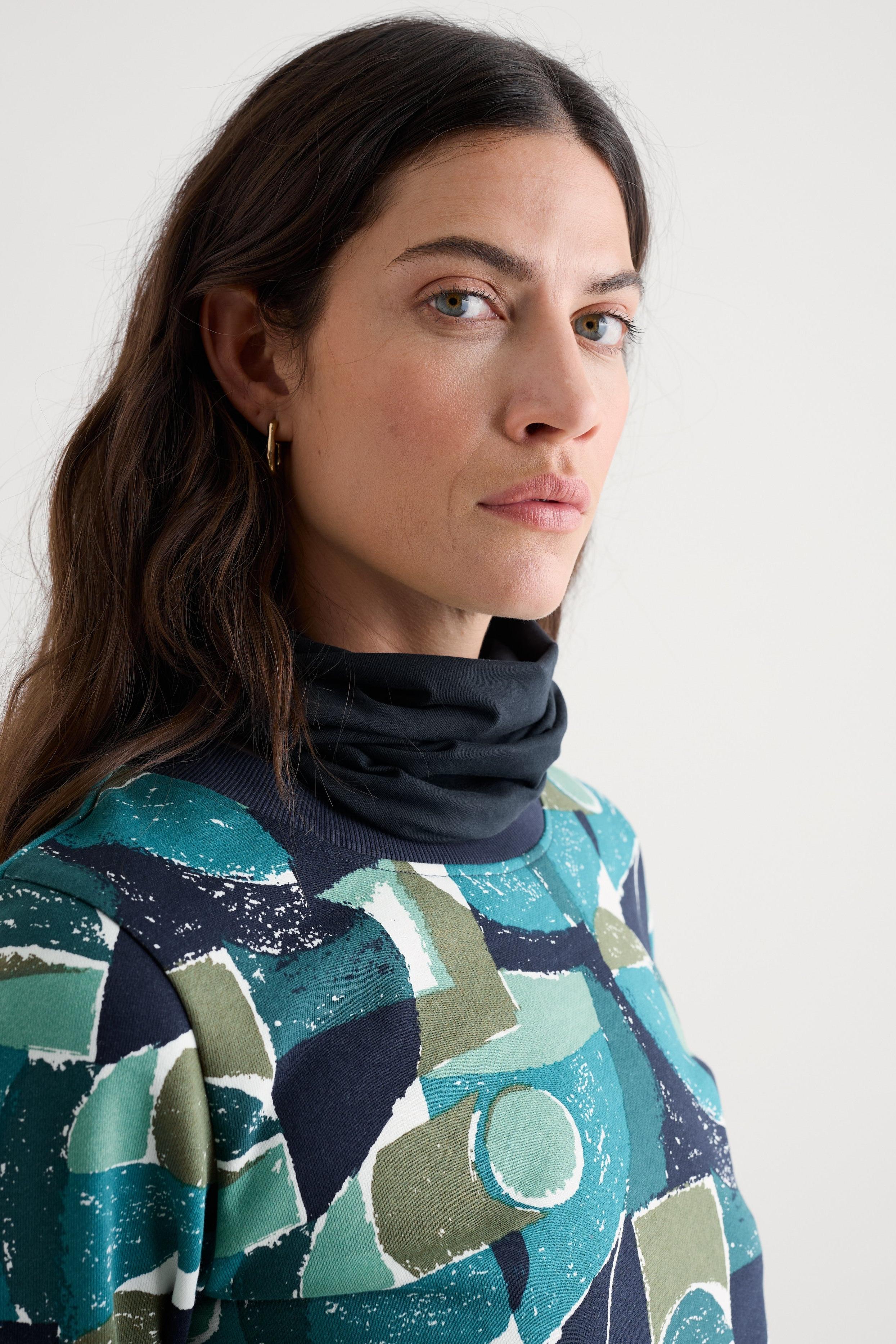 A woman wearing a dark navy infinity scarf with an abstract print knit top in shades of teal, green, and navy, photographed against a plain light gray background.
