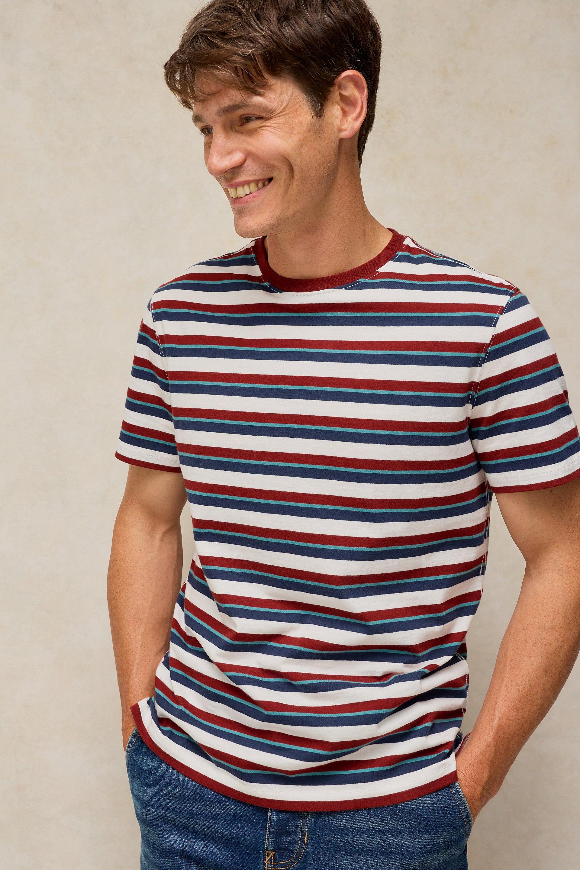 A man wearing a short-sleeve crewneck t-shirt with horizontal red, navy blue, and white stripes, paired with blue jeans, set against a neutral textured beige background.