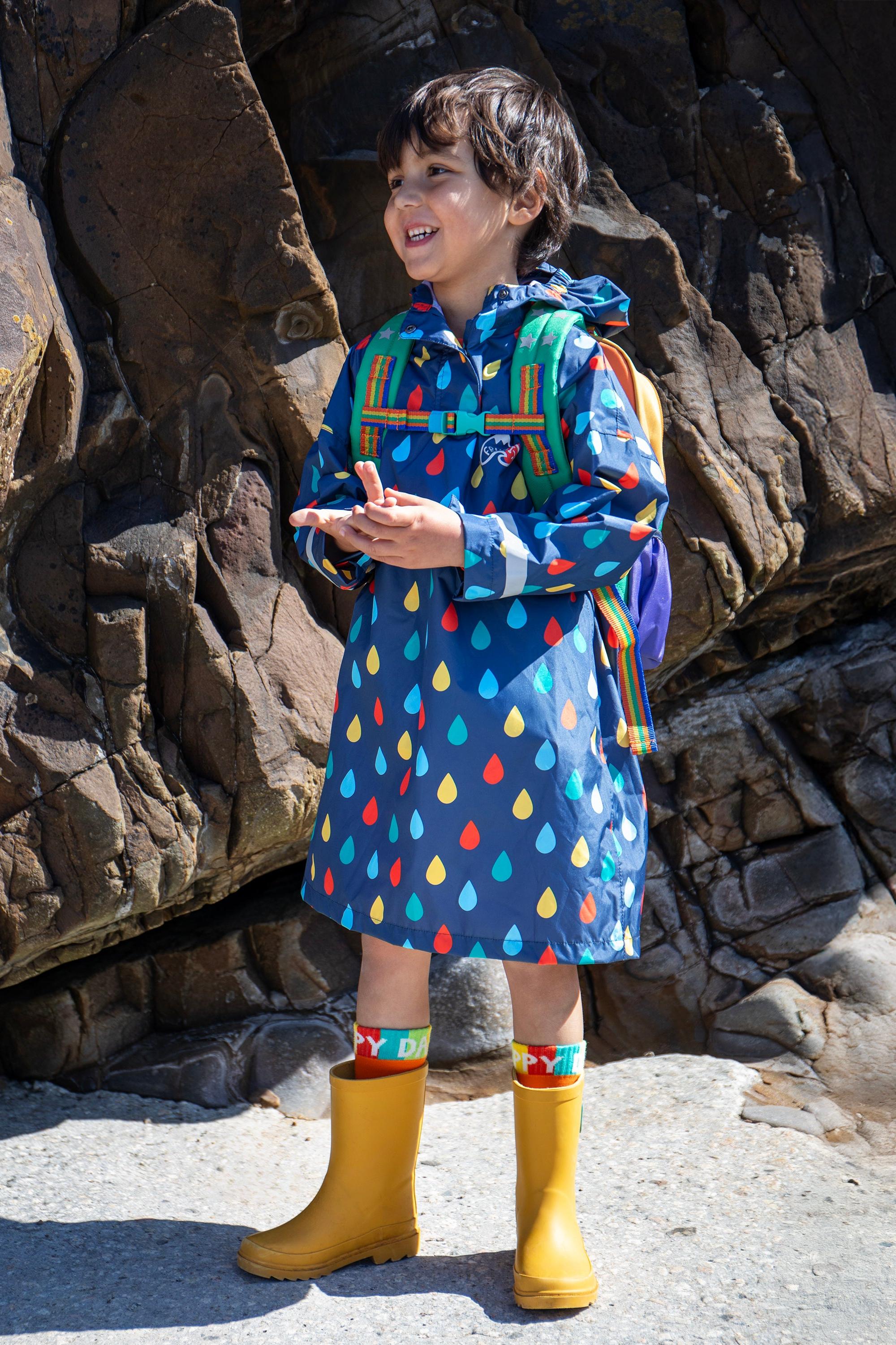 Child wearing a colorful Frugi raincoat with a rainbow pattern, standing against a rocky background.
