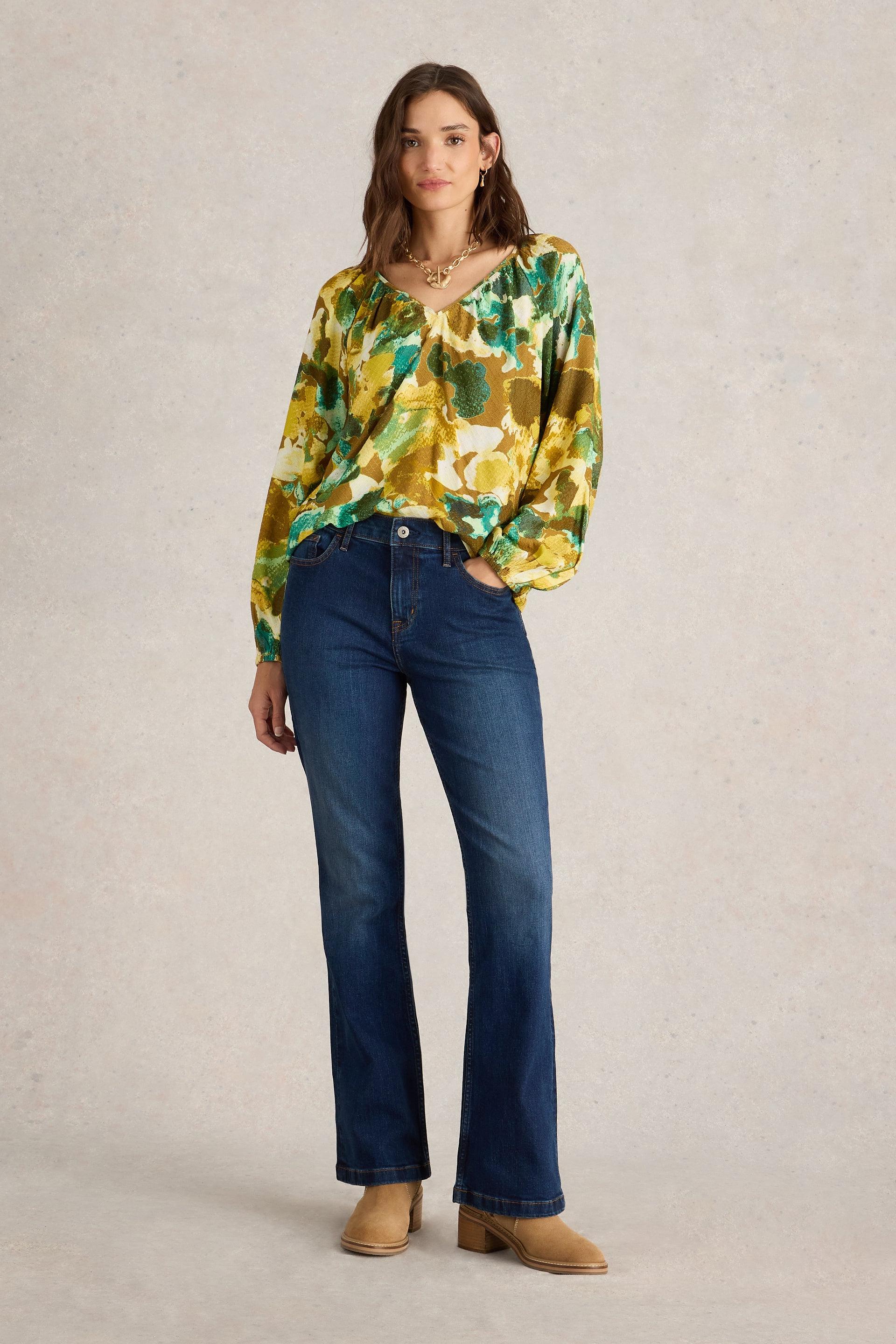 A woman modeling a colorful floral blouse and blue bootcut jeans against a neutral studio background.