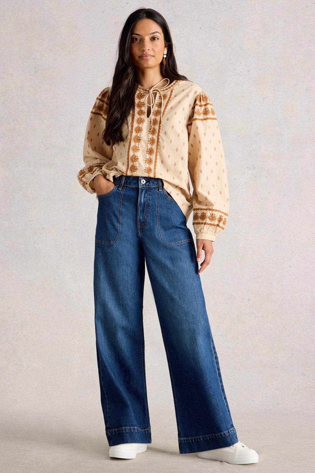 A woman modeling a beige patterned long-sleeve blouse with a tie neckline, tucked into high-waisted wide-leg blue jeans, against a textured light gray studio backdrop.