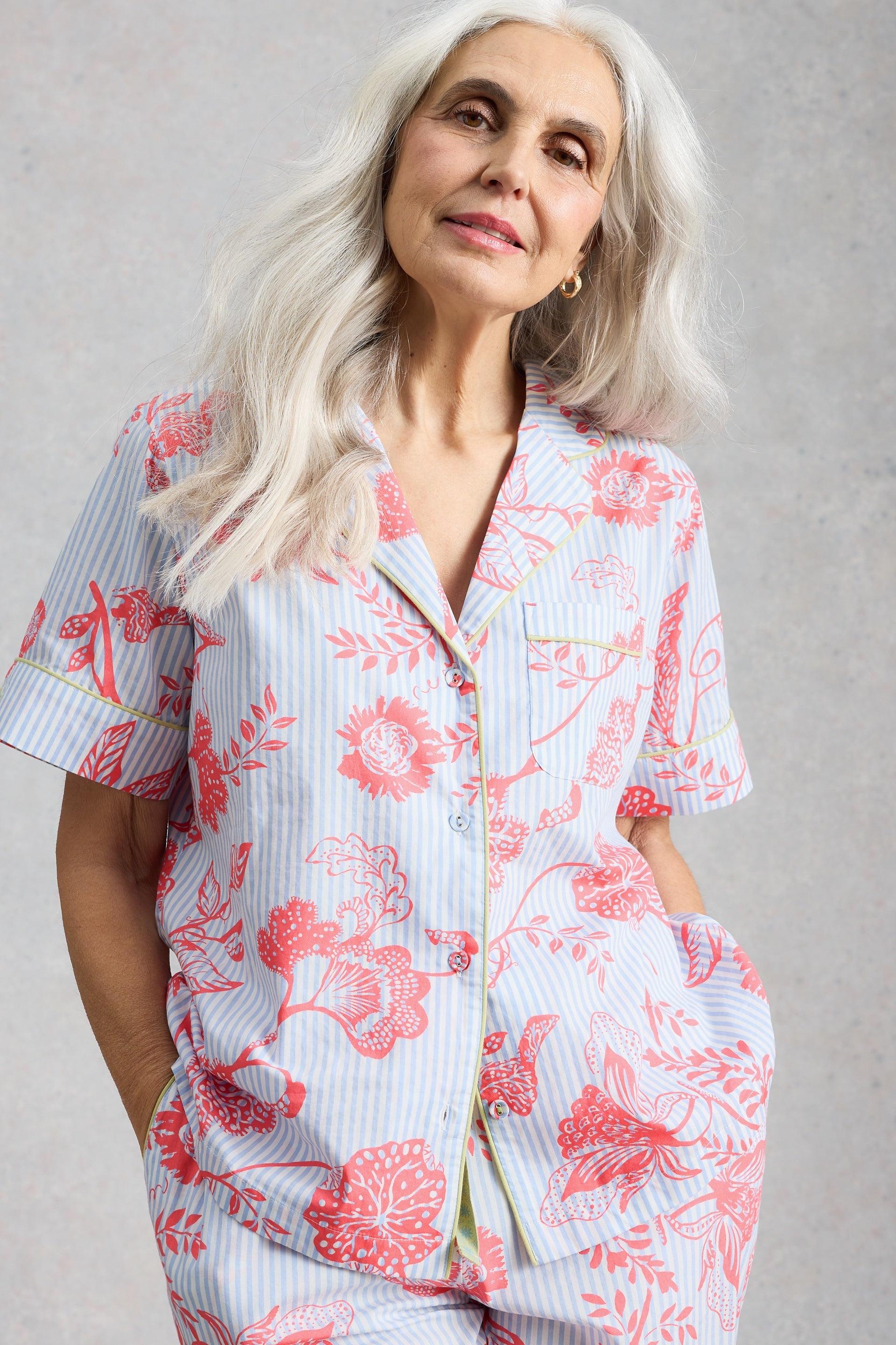 A smiling mature woman with long grey hair wearing blue and red floral print organic cotton pyjamas.