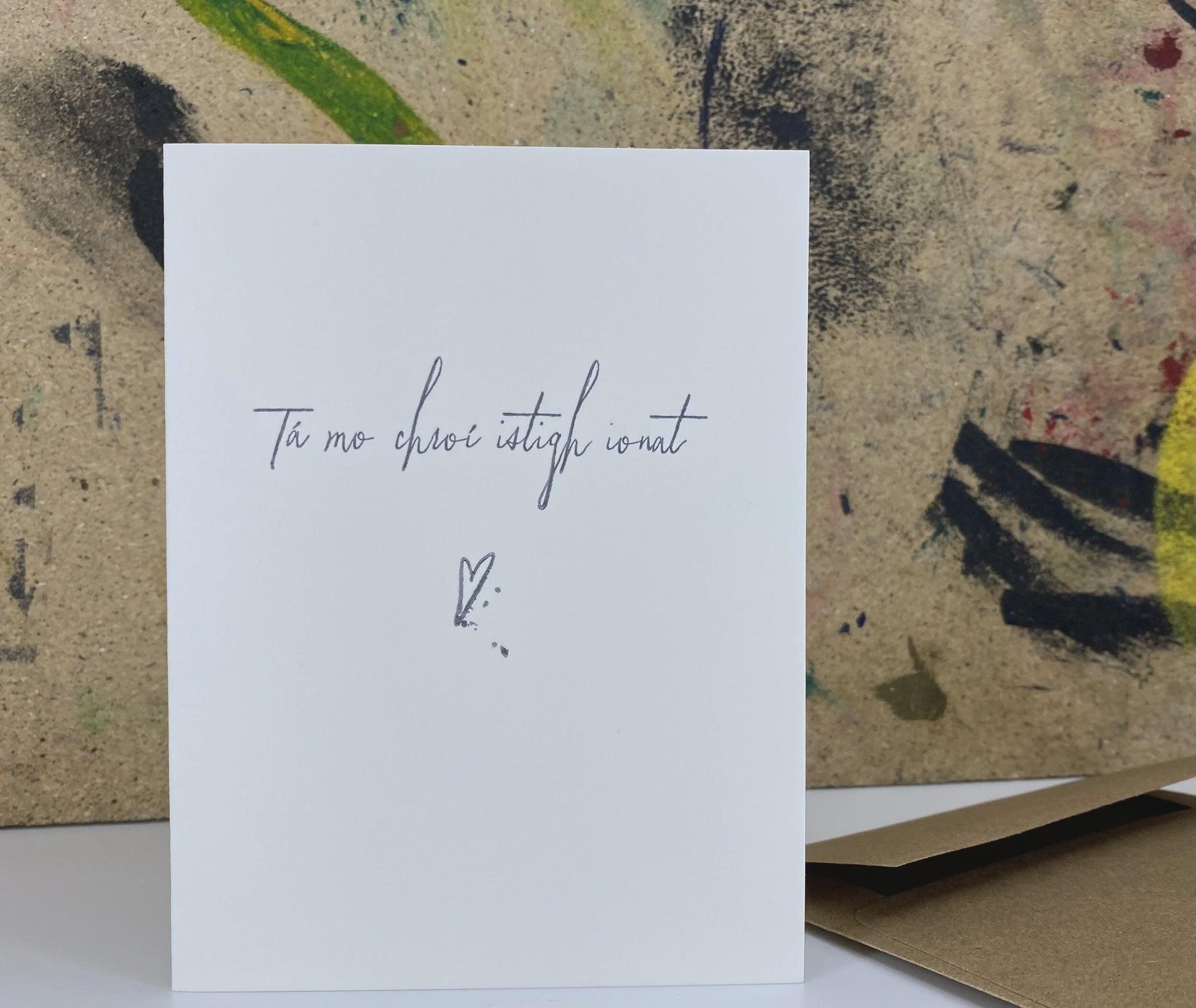 A white greeting card with elegant Irish text reading 'Tá mo chroí istigh ionat' and a small heart with a dot below, displayed in front of a textured, abstractly painted background.