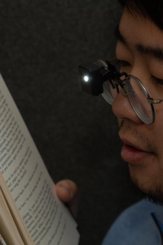 A close-up side profile of a man reading an open book while wearing a black magnifying glass attachment clipped to his metal-framed eyeglasses. The small built-in light is turned on, illuminating the book pages against a dark background.