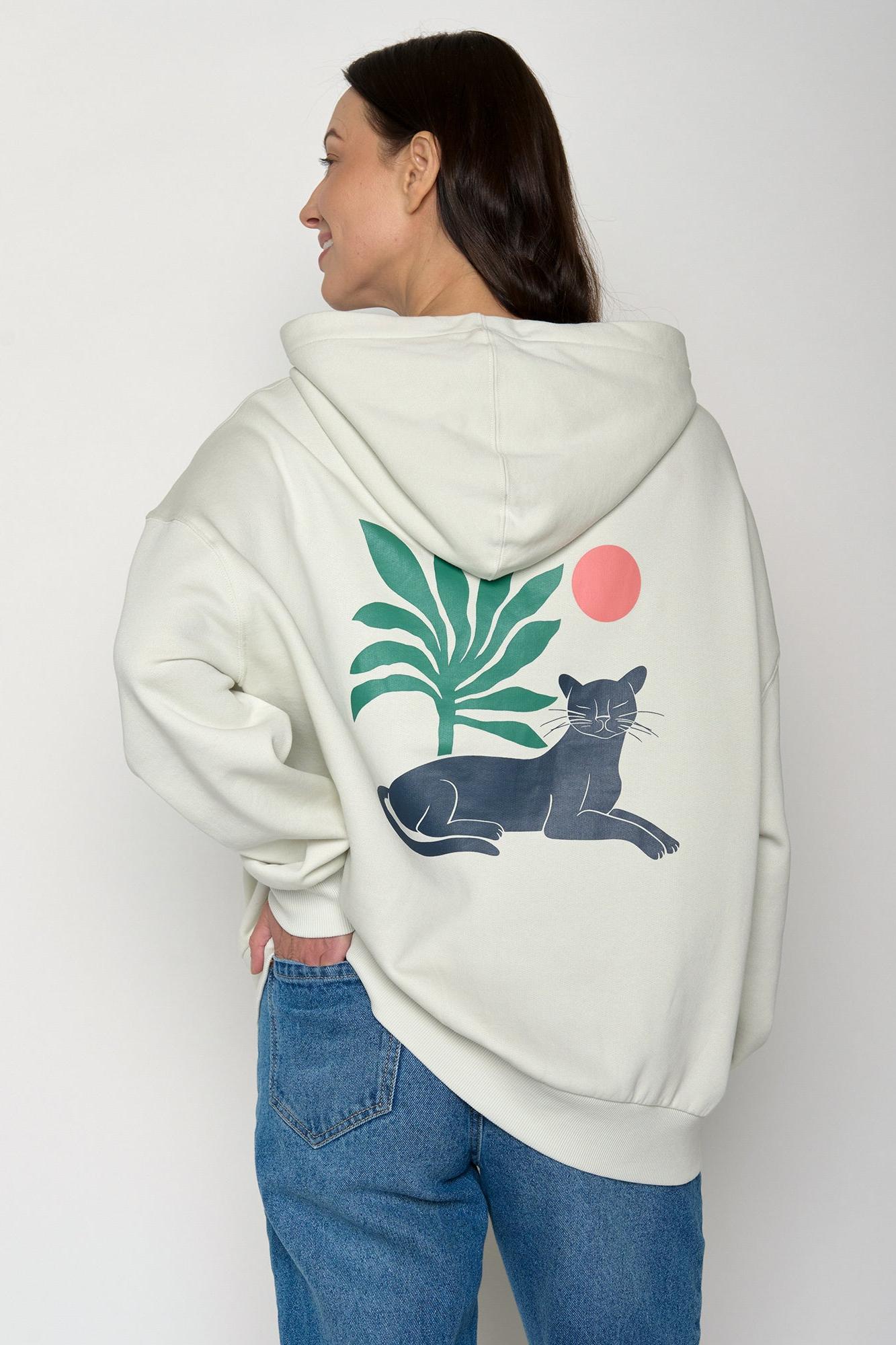 Person wearing a cream hoodie with a graphic of a panther, palm leaves, and a red circle on the back.