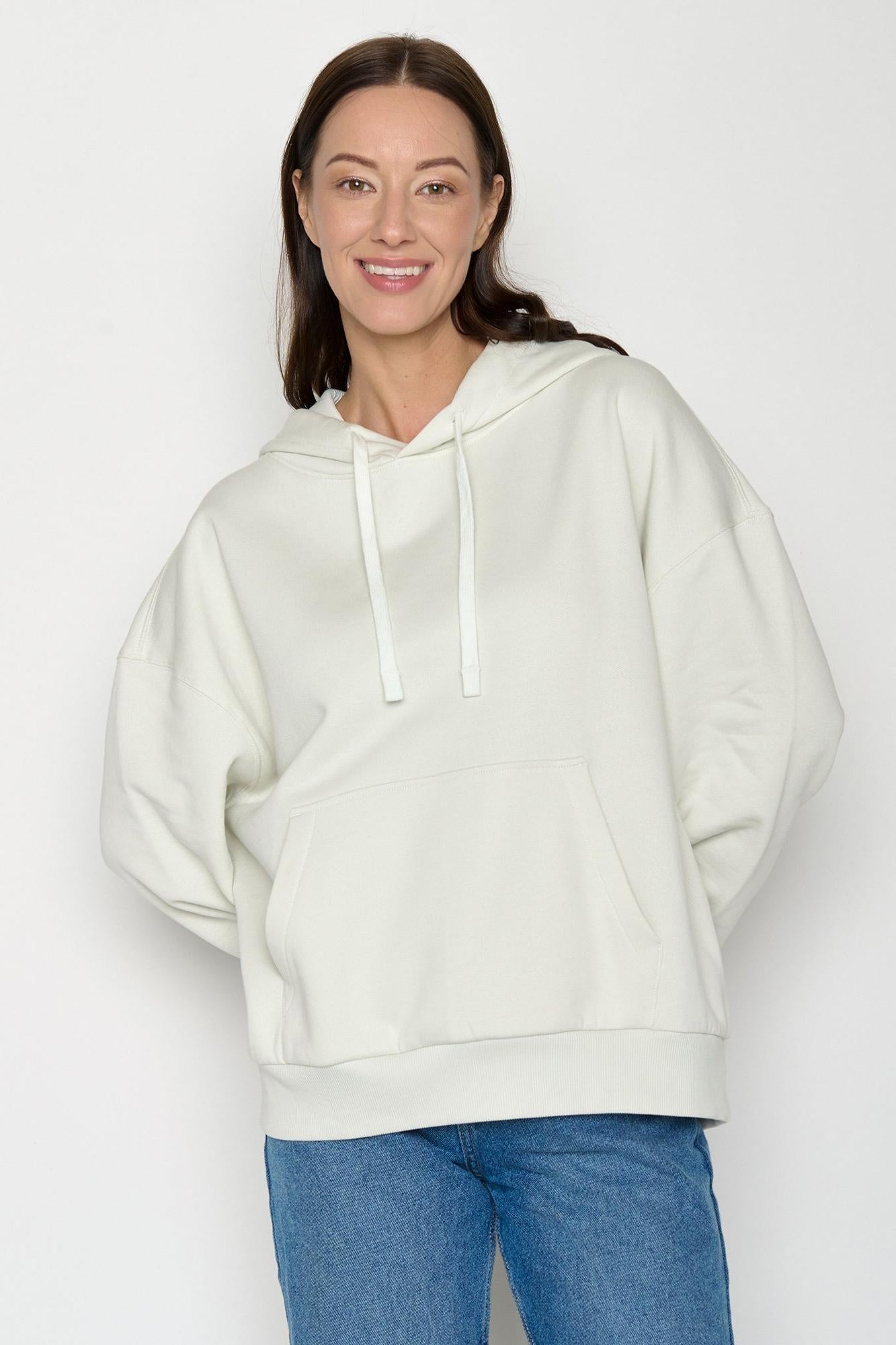 Woman wearing a white organic cotton hoodie and blue jeans against a plain background