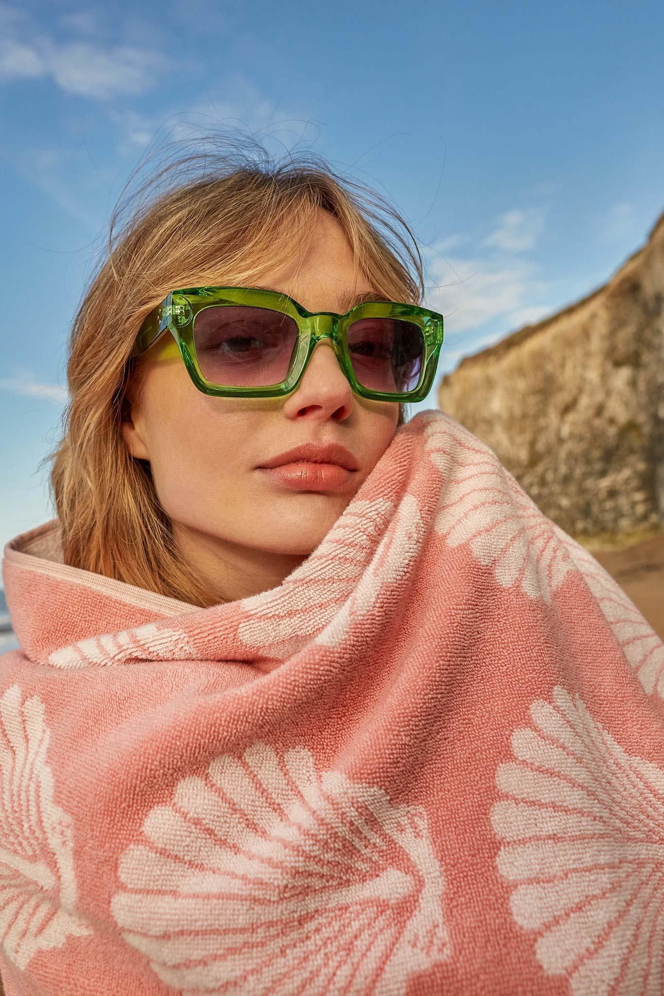 Woman wearing green sunglasses and a pink patterned towel on a beach.
