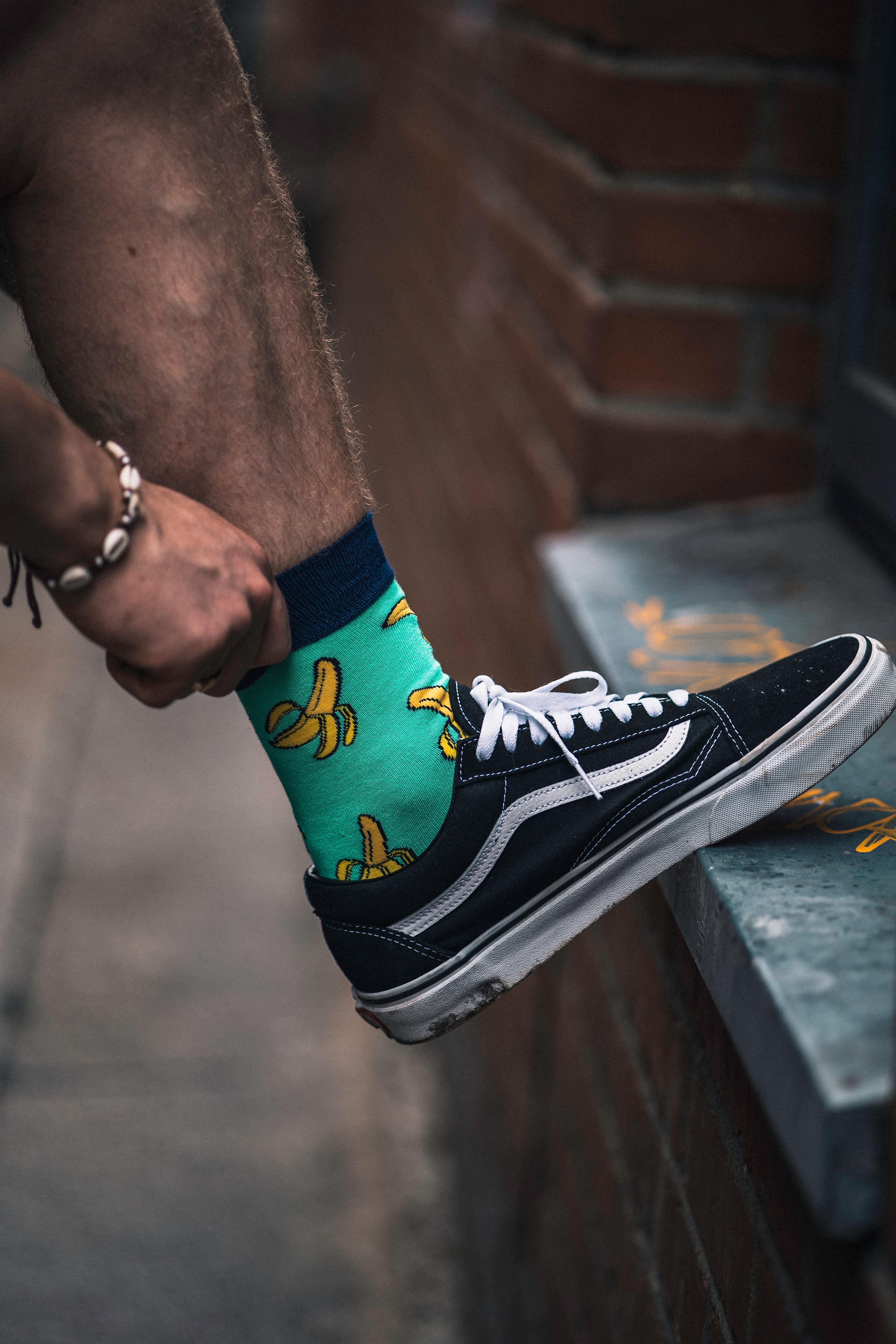 Person wearing a black and white sneaker with a green bamboo sock featuring banana prints, standing on a concrete surface.
