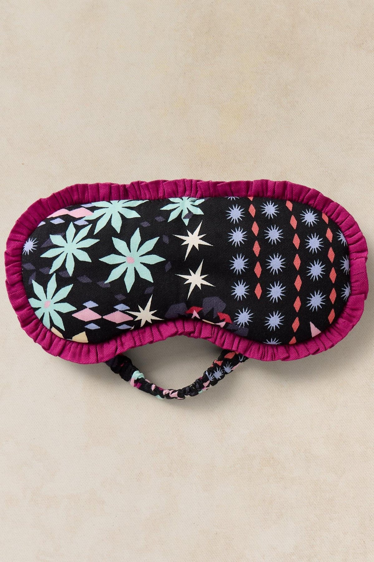 A fabric sleep mask with a black background featuring a colorful star and floral pattern, finished with a magenta ruffled edge and an elastic strap, shown on a textured beige background.
