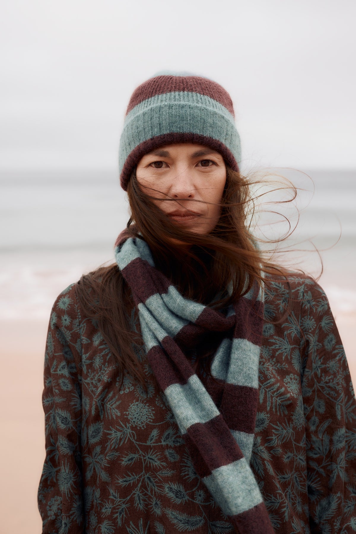 A woman standing outdoors on a beach with the wind blowing her hair, wearing a striped knit beanie in dusty teal and burgundy, a matching striped knit scarf, and a dark brown patterned coat.