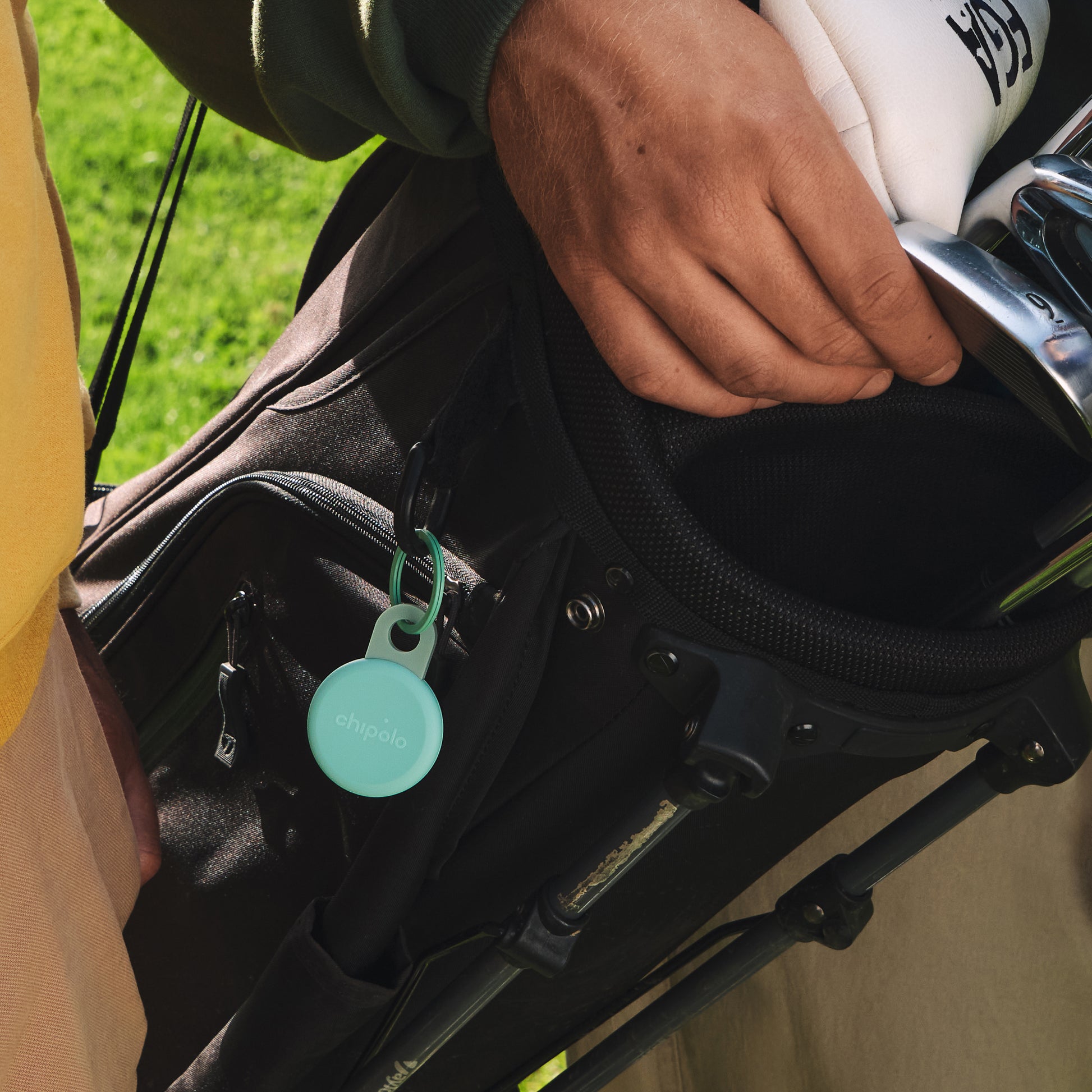 Close-up of a golf bag with a hand holding a club, featuring a turquoise Chipolo tracker tag.