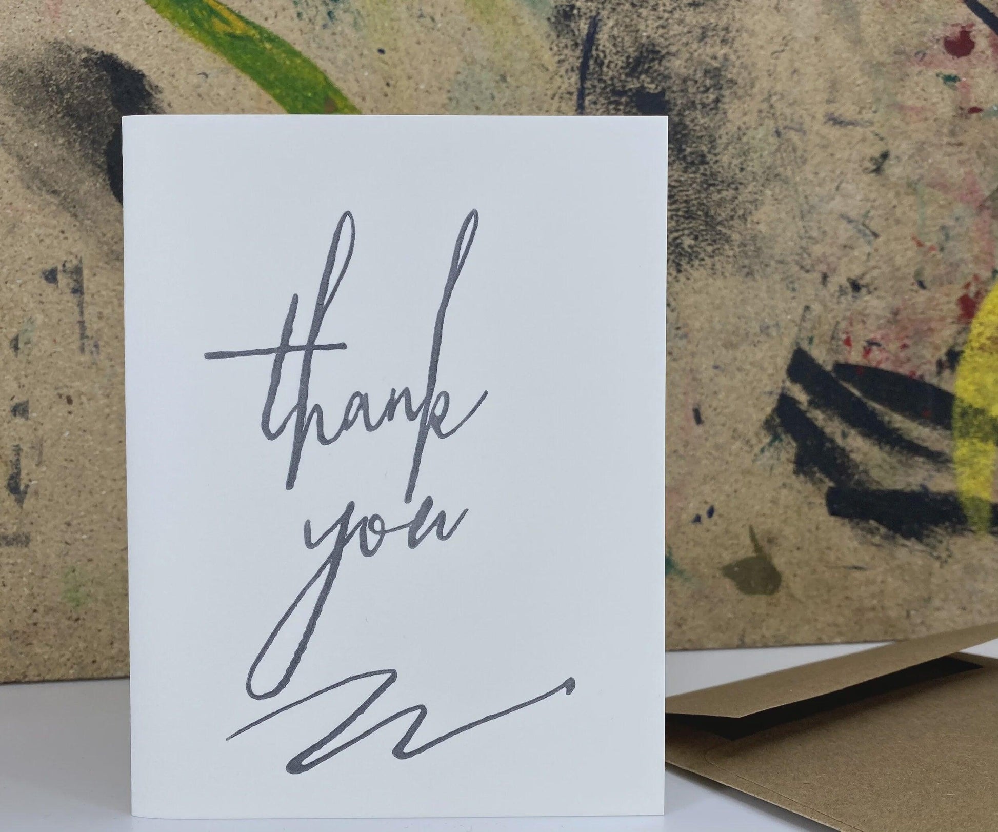 A white square greeting card with the words 'thank you' written in gray script, standing upright in front of a textured abstract painting. A brown envelope is partially visible at the lower right.
