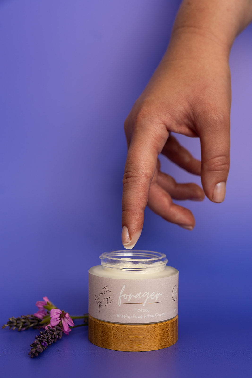 Hand reaching towards a forager body and eye cream labeled 'forager' on a purple background