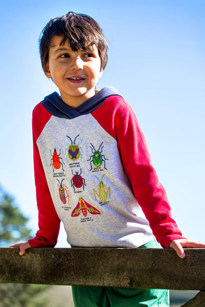Child leaning on fence wearing GOTS organic cotton hooded top with bug design, red sleeves and navy hood