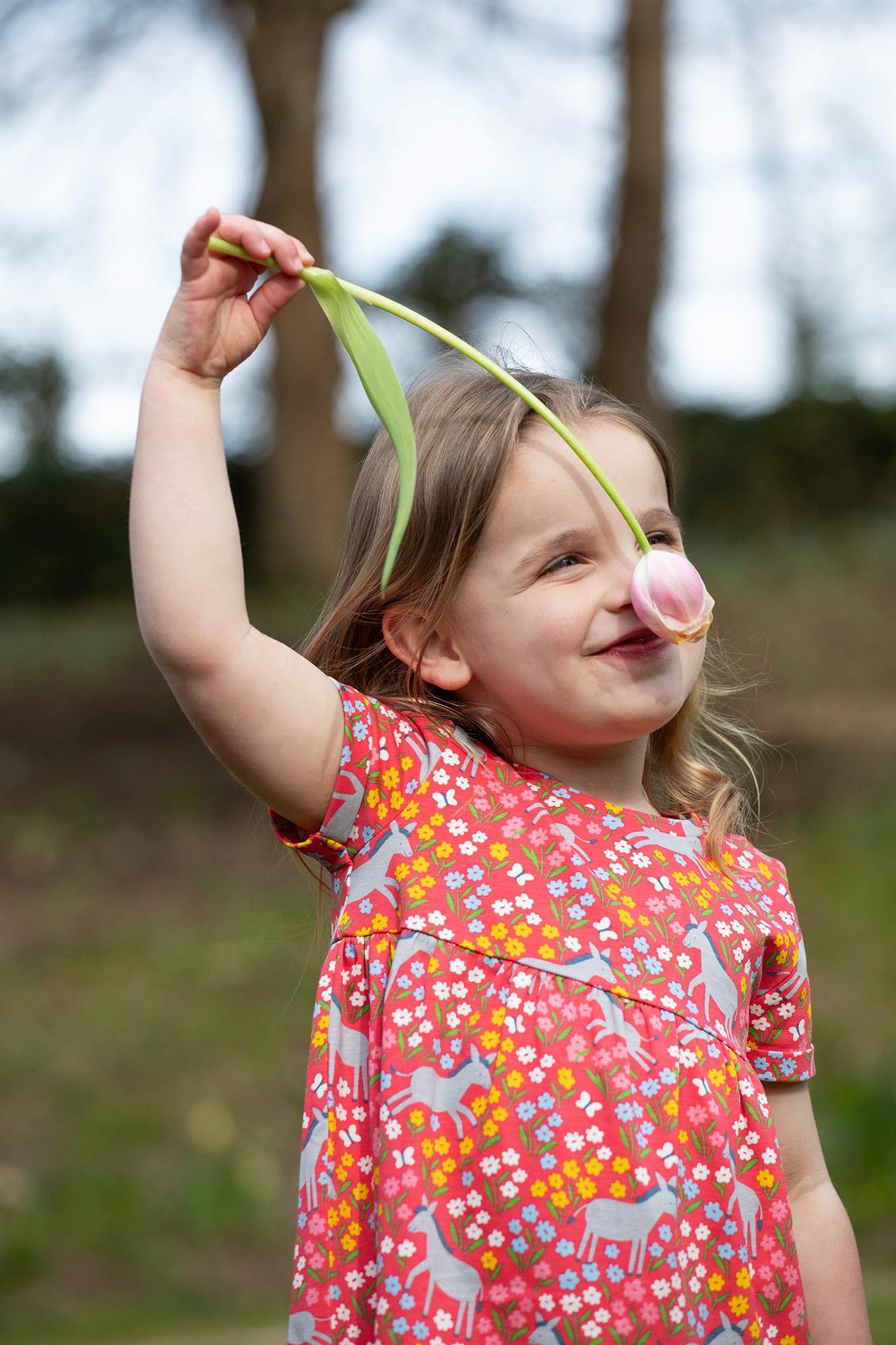 Young girl wearing a red short-sleeved dress with a playful donkey print, smiling and smelling a flower.