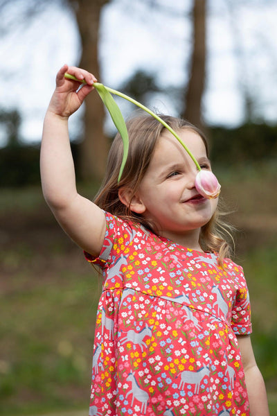 Young girl wearing a red short-sleeved dress with a playful donkey print, smiling and smelling a flower.