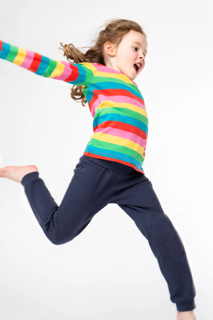 Child wearing a colourful striped shirt and organic cotton navy pants jumping on a white background