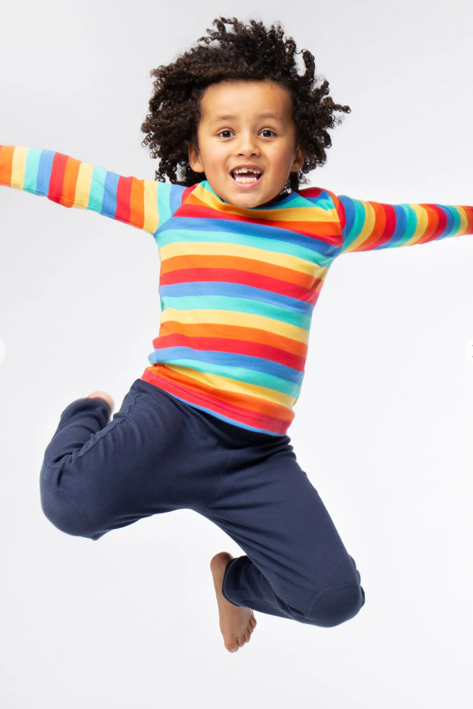 Child wearing a colourful striped shirt and blue organic cotton pants jumping against a white background