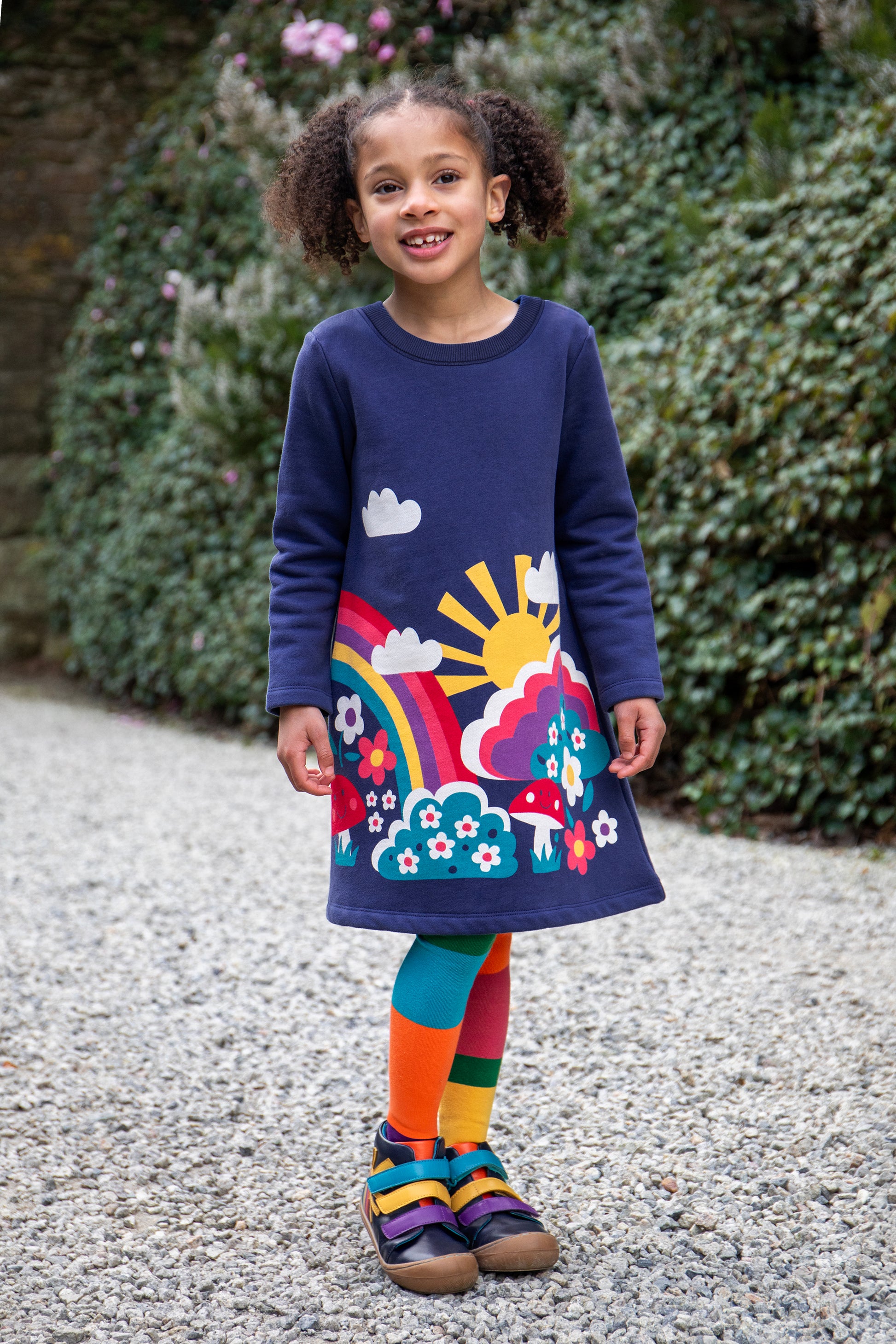Child wearing a colorful organic cotton dress with a rainbow and floral design, standing outdoors.