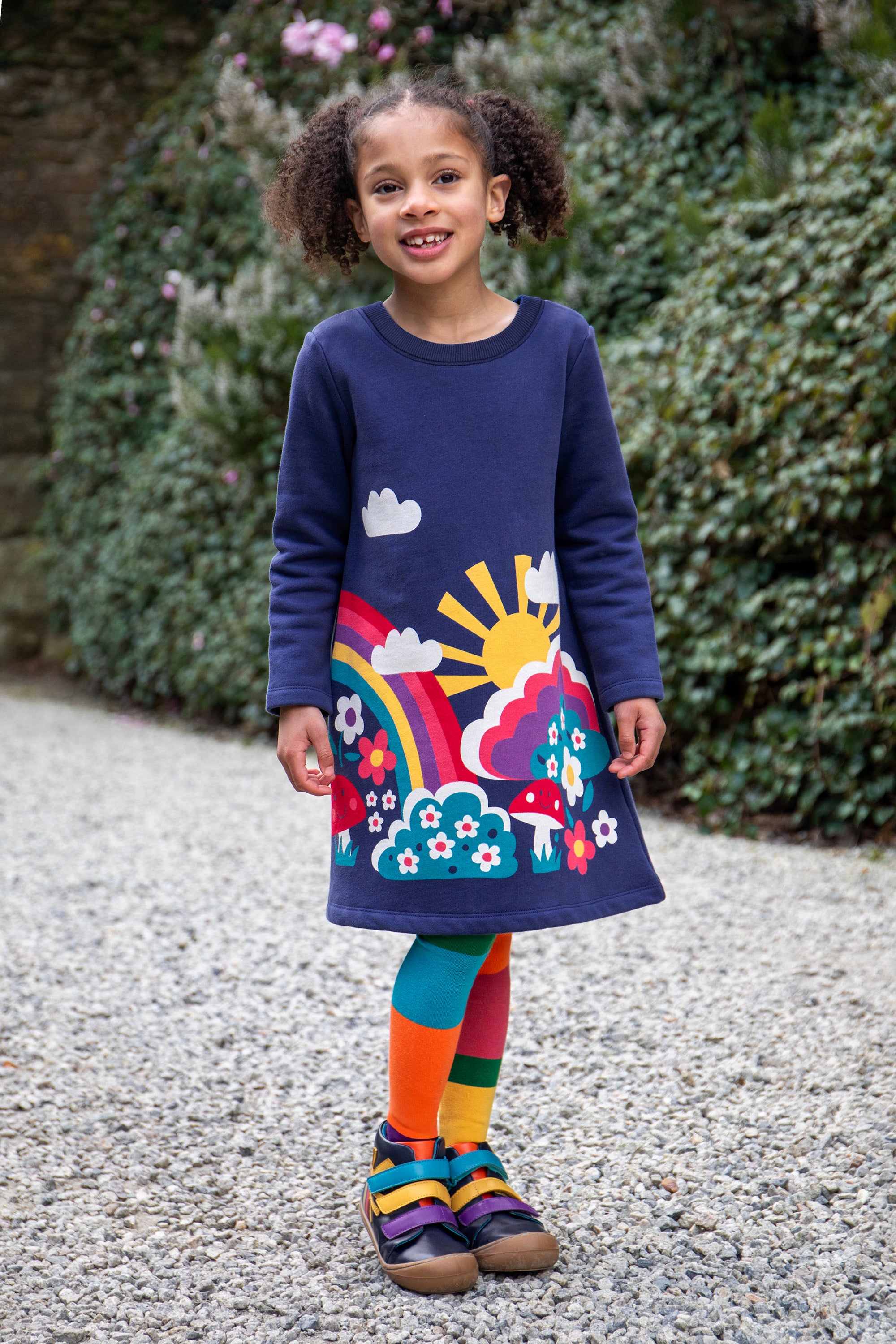 Child wearing a colorful organic cotton dress with a rainbow and floral design, standing outdoors.