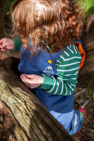 Child climbing outdoors in Frugi navy waterproof dungarees layered over green striped top