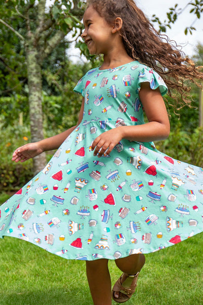 Young girl wearing a blue short-sleeved skater dress with colourful baking-themed illustrations, smiling and looking to the side.