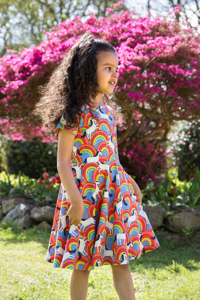Child wearing a colourful rainbow-print dress with short sleeves, smiling outdoors.