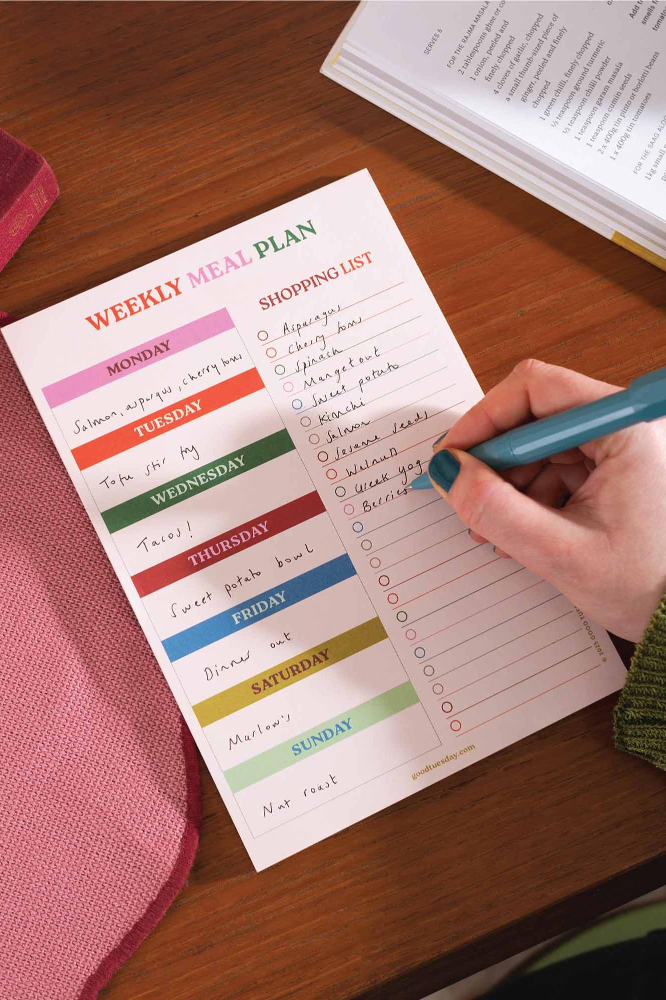 Weekly meal plan and shopping list on a wooden table with a hand holding a pen.