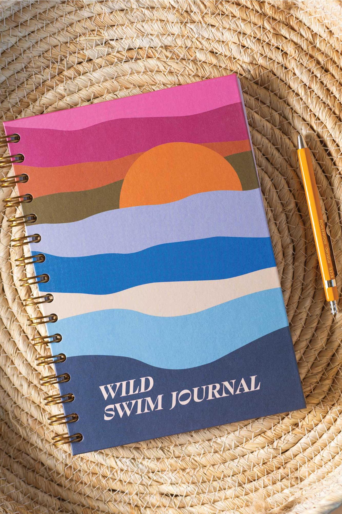 Notebook titled 'Wild Swim Journal' with a sunset design on a woven surface