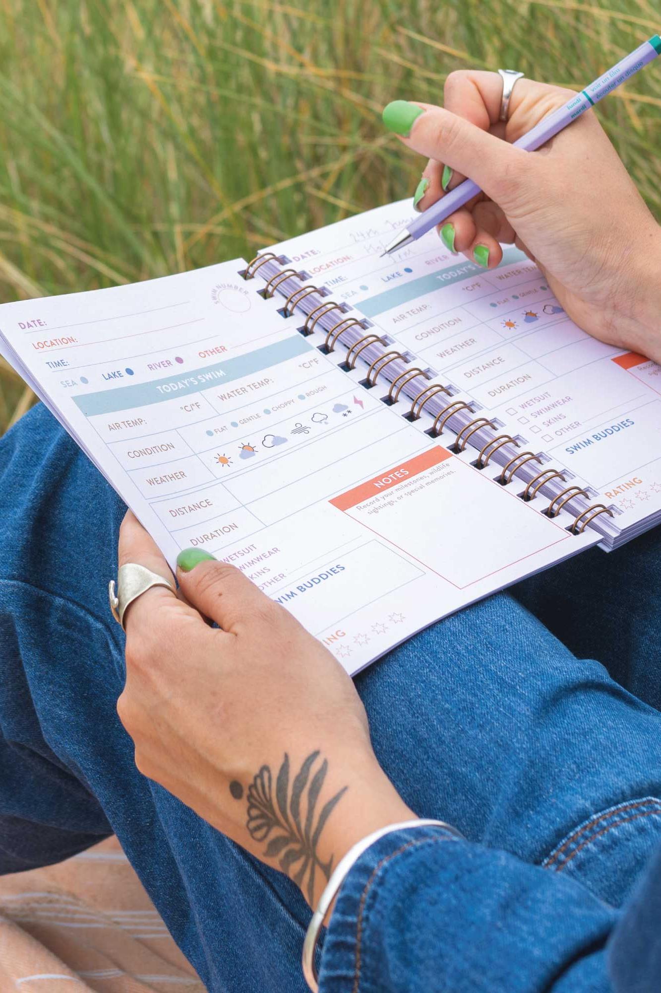 Person holding a planner with a pen, sitting outdoors on grass.