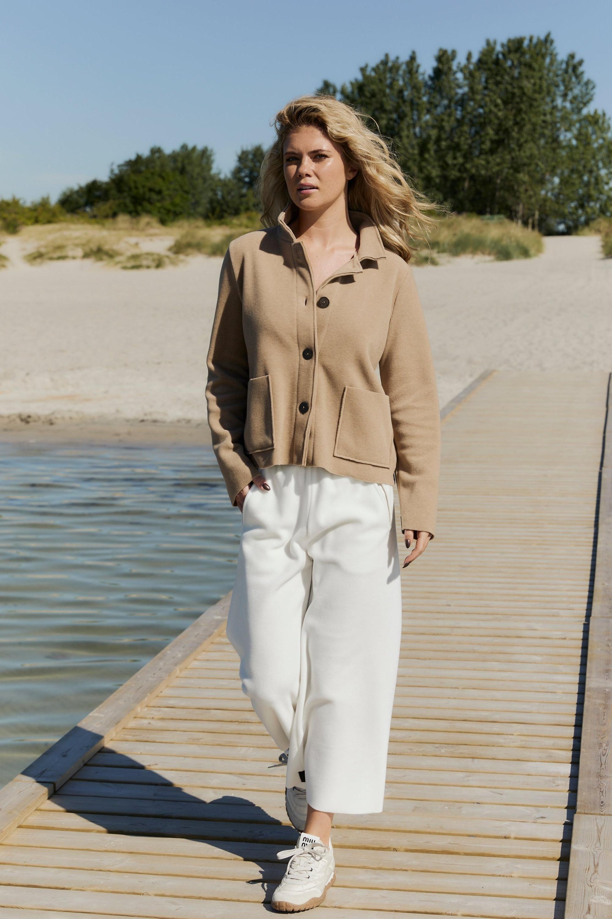 Woman standing on a wooden dock by a body of water, wearing a beige fleece jacket and white pants.