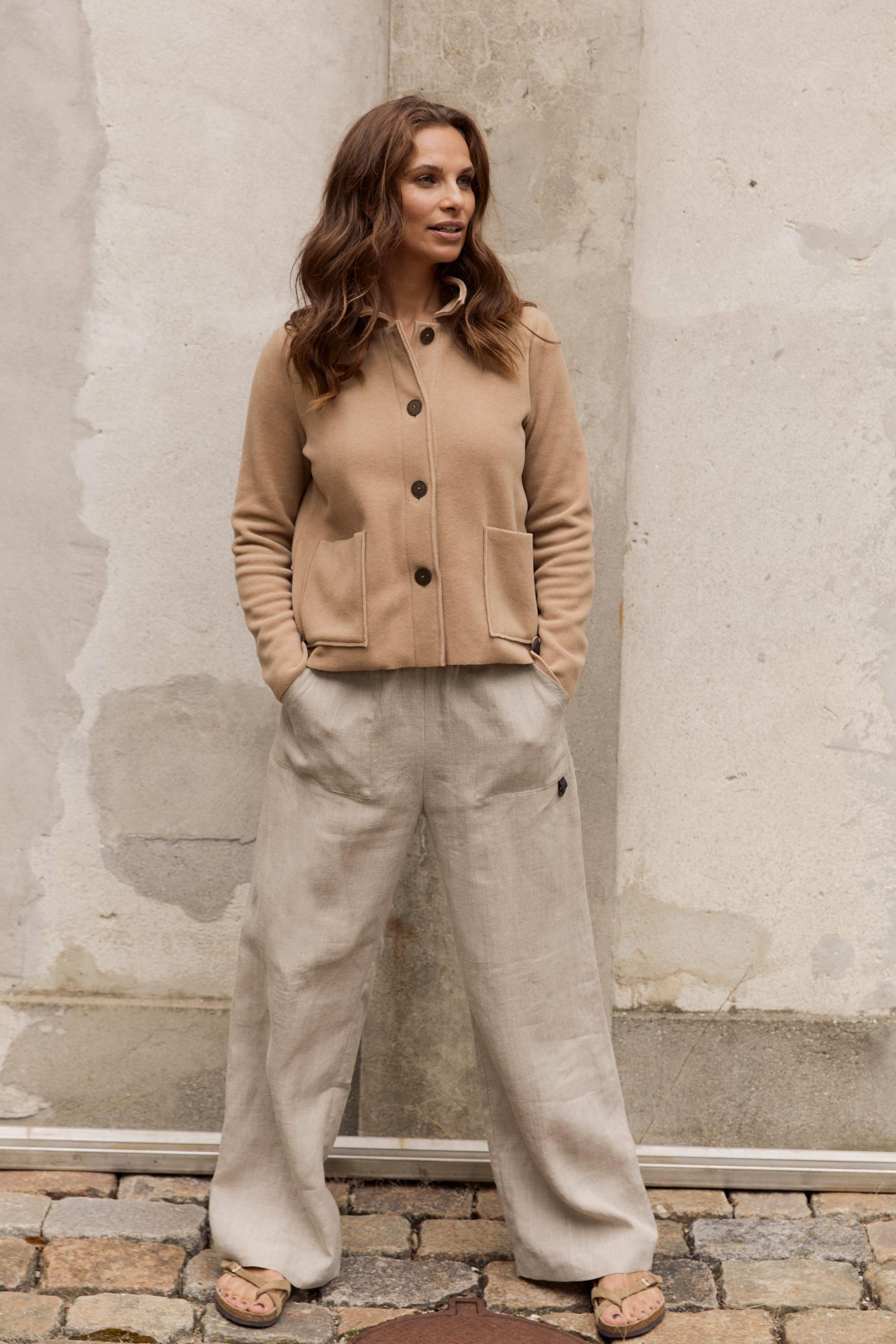 Woman wearing a beige fleece jacket and light gray pants standing against a textured wall.