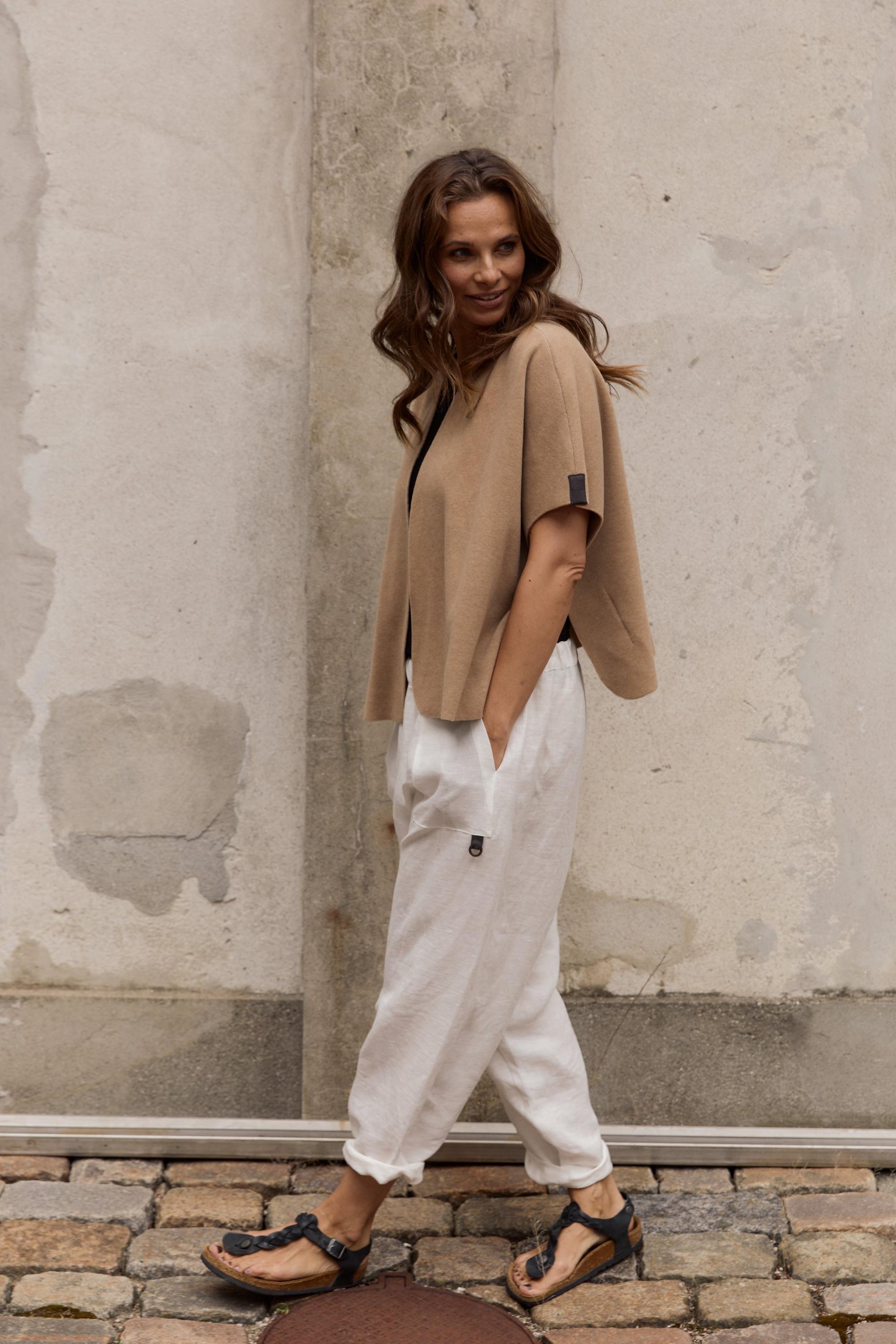 Woman wearing a beige fleece top and white pants standing against a textured wall.