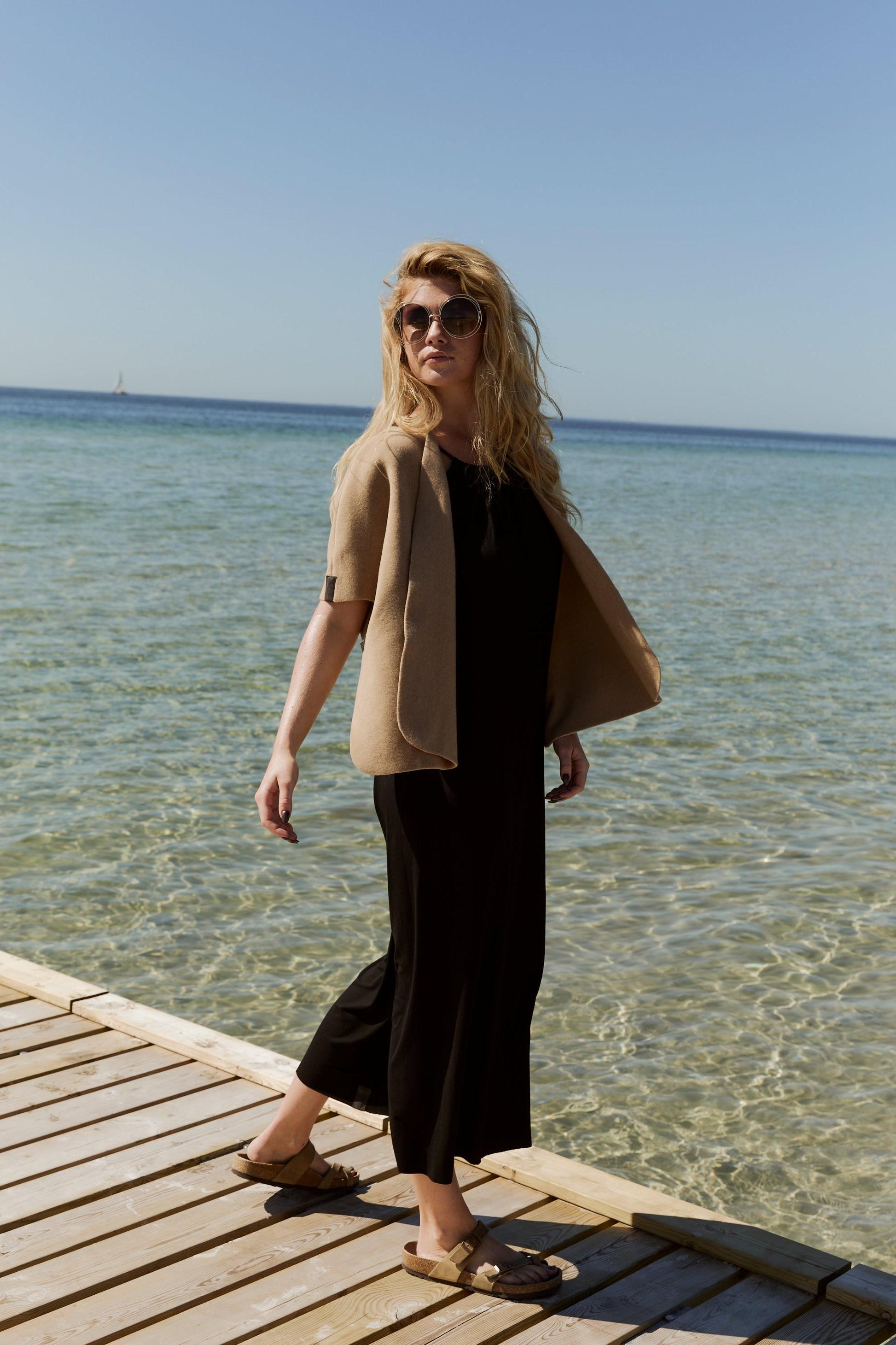 Woman standing on a wooden dock by the water wearing sunglasses and a beige fleece cardigan.