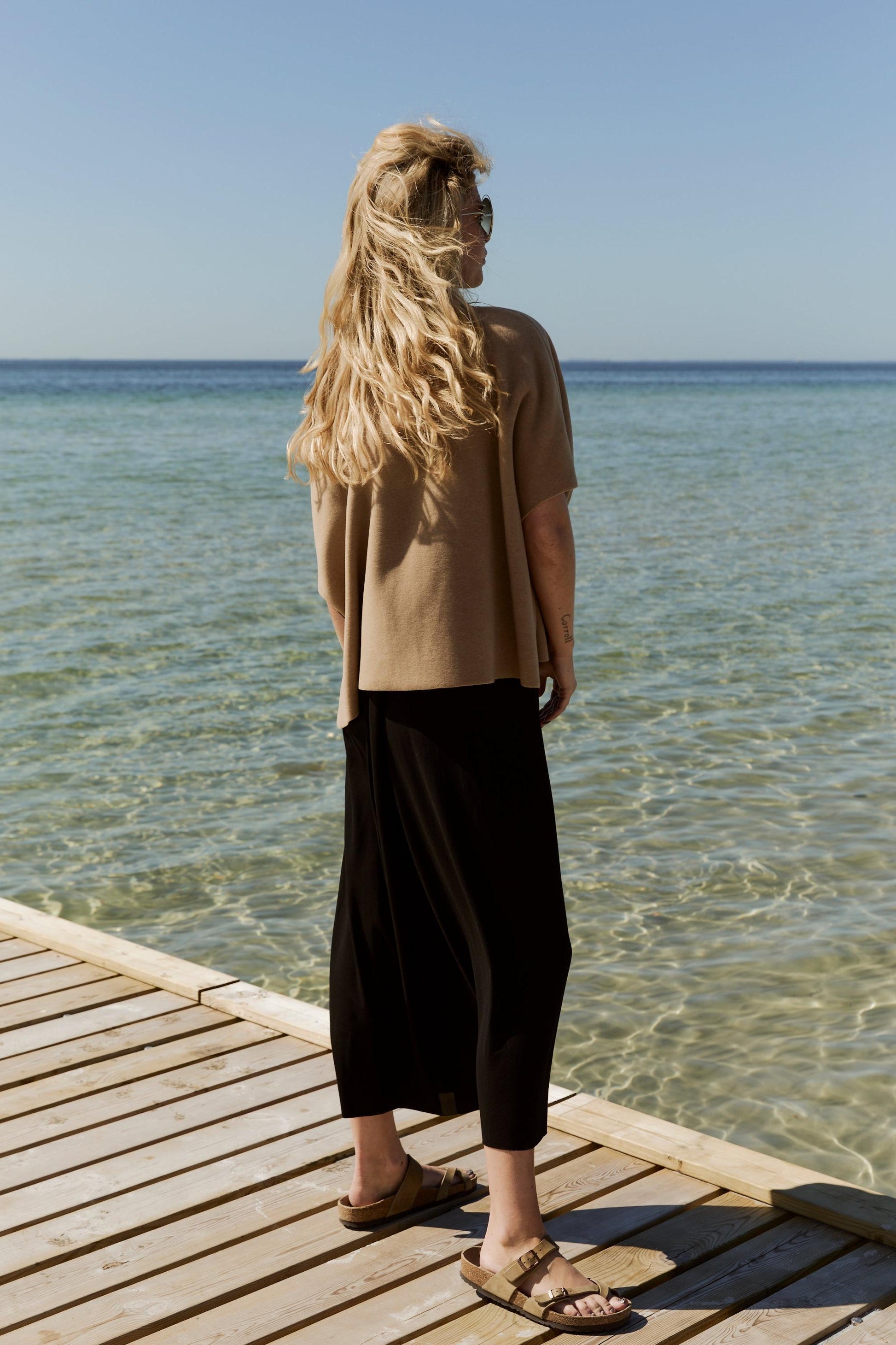 Woman standing on a wooden dock by the water wearing a beige fleece top and black pants.