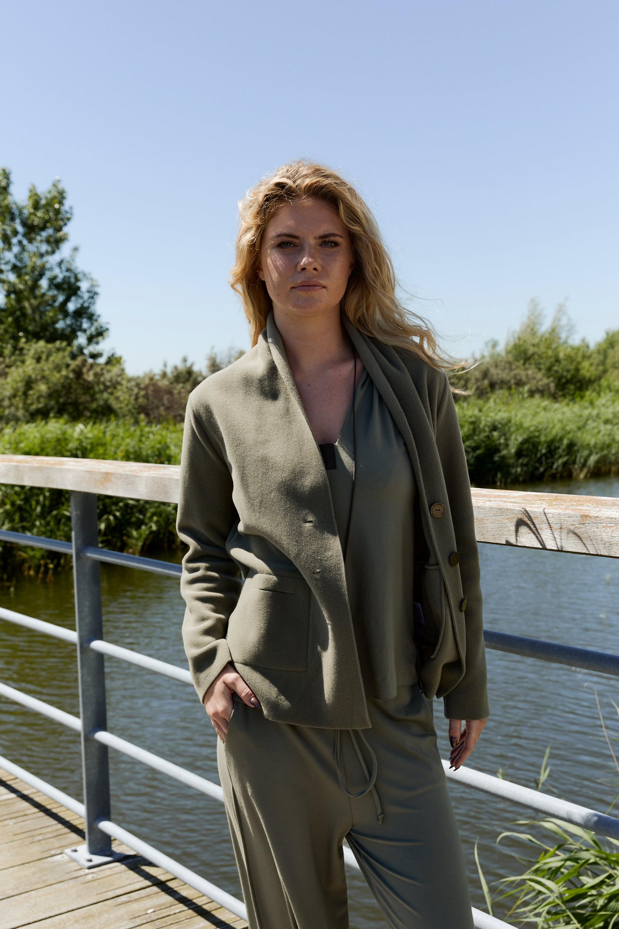 Woman in a green fleece cardigan standing on a wooden bridge by a body of water.