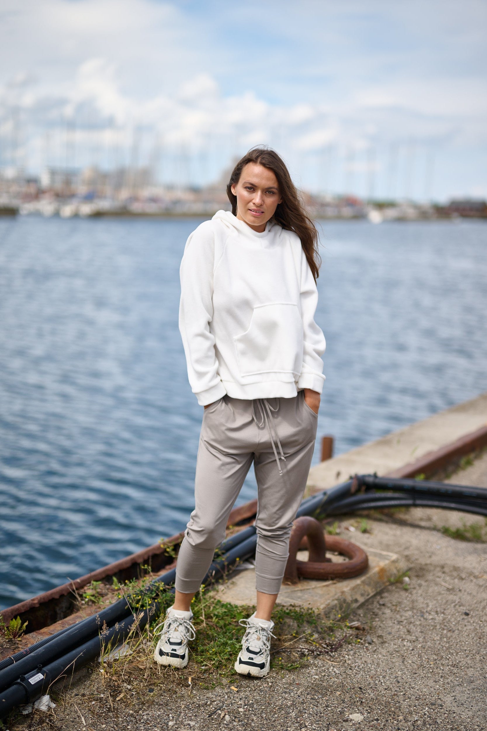 Model standing on a pier, wearing a white hoodie paired with grey lounge pants and white trainers.