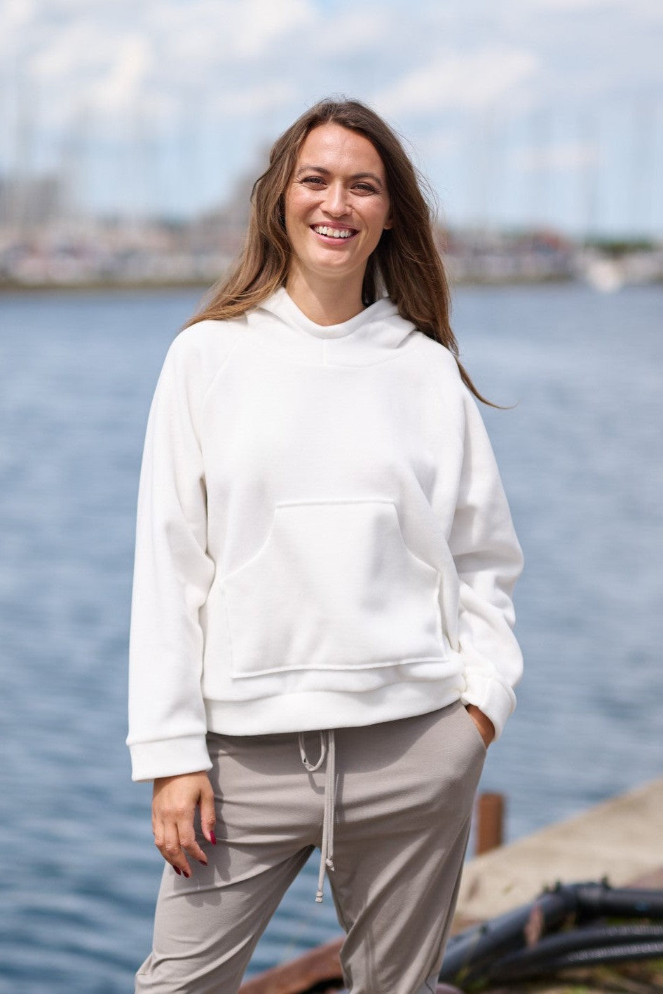 Model at a waterfront scene, wearing a white hoodie featuring raglan sleeves and a front pocket.