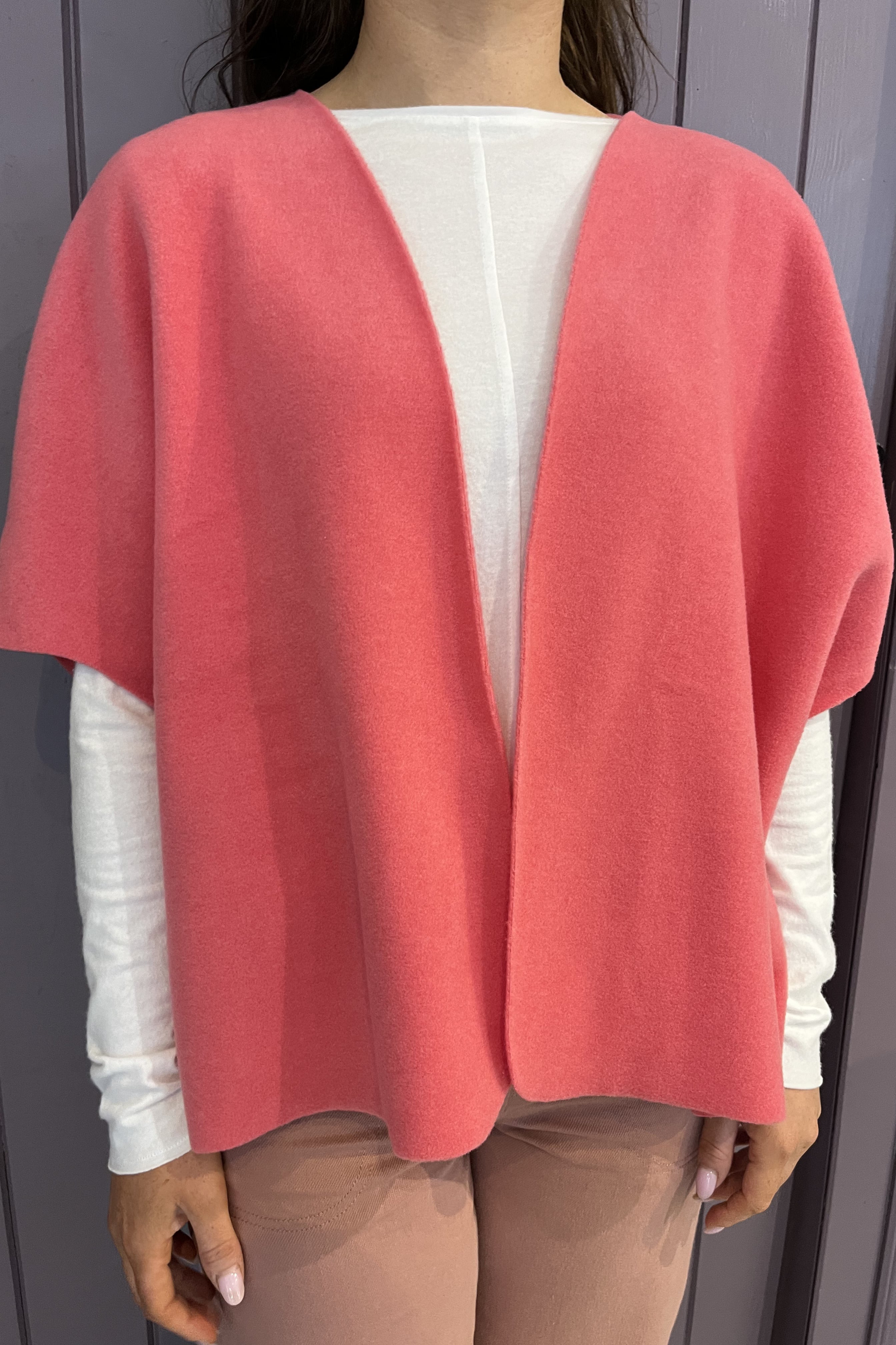 A woman wearing a coral pink open-front poncho-style cardigan over a white long-sleeve top and beige pants, shown from the shoulders to the upper thighs against a gray paneled background.