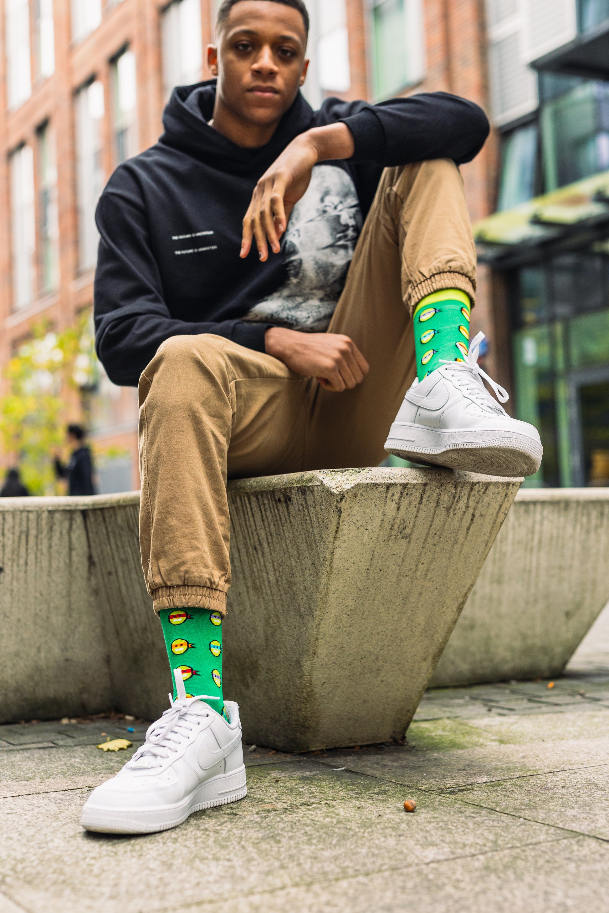 Person sitting on a concrete bench wearing a black hoodie, beige pants, white sneakers, and colourful bamboo socks.