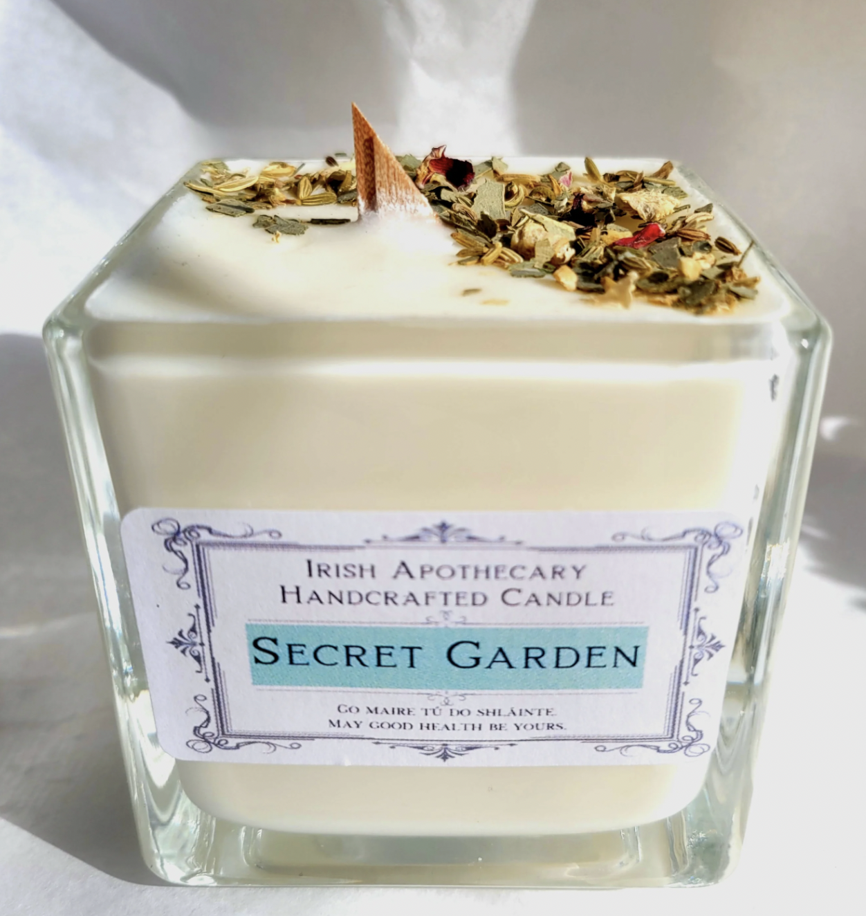 Square glass candle labeled 'Secret Garden' with dried herbs on top, on a white background.