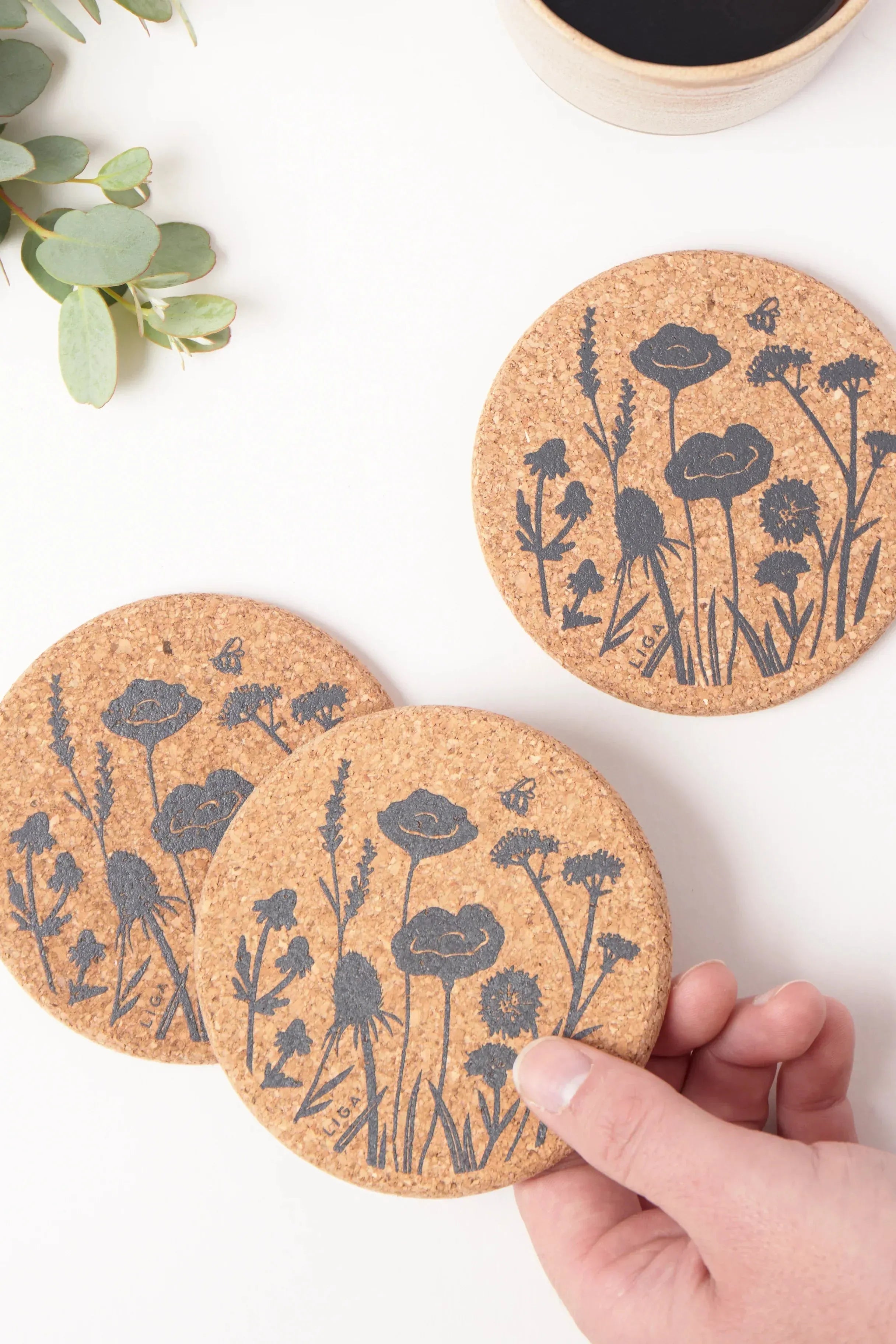 Cork coasters with floral designs held by a hand on a white surface with a cup and plant.
