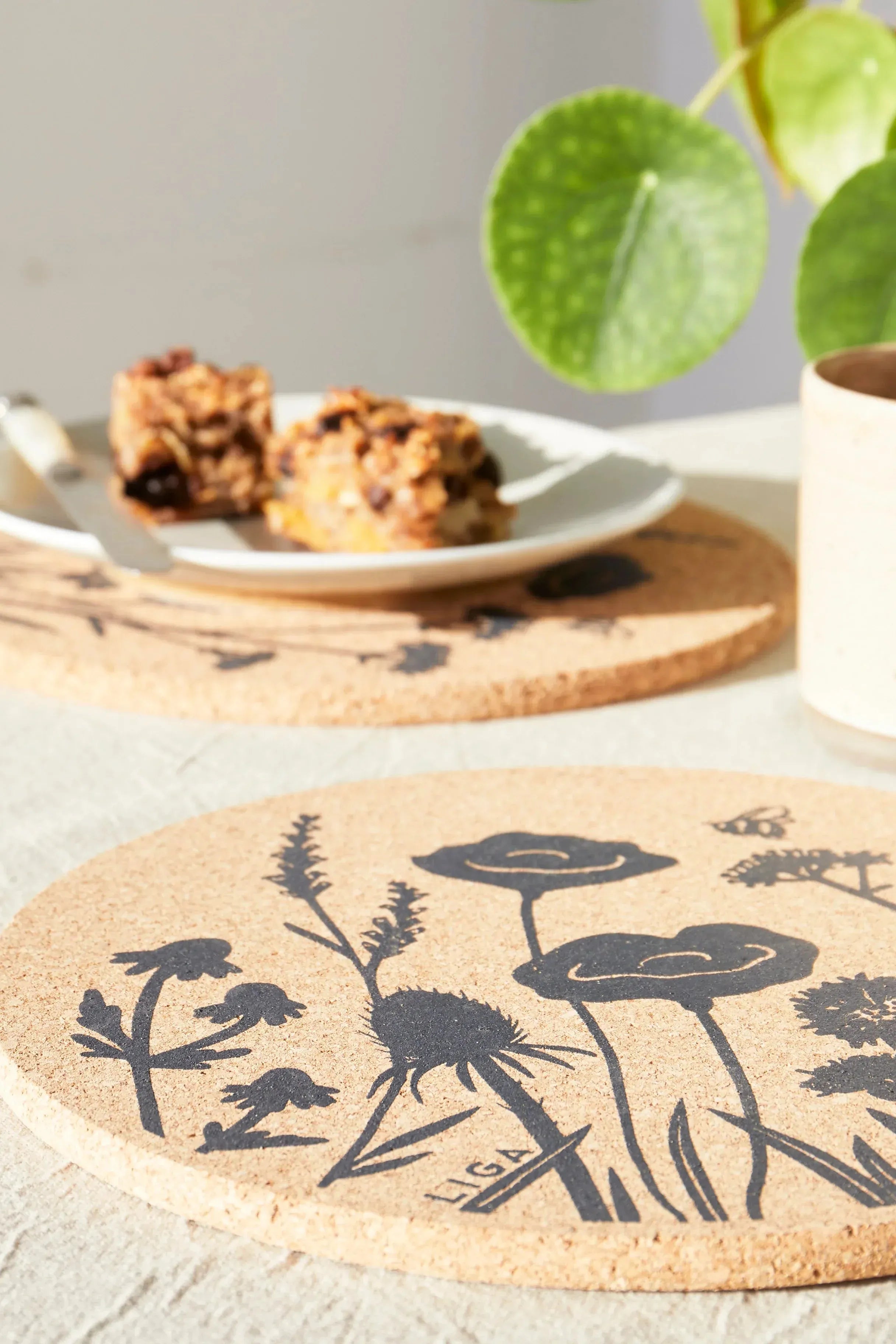 Cork coaster with floral design on a table with cookies and a plant.