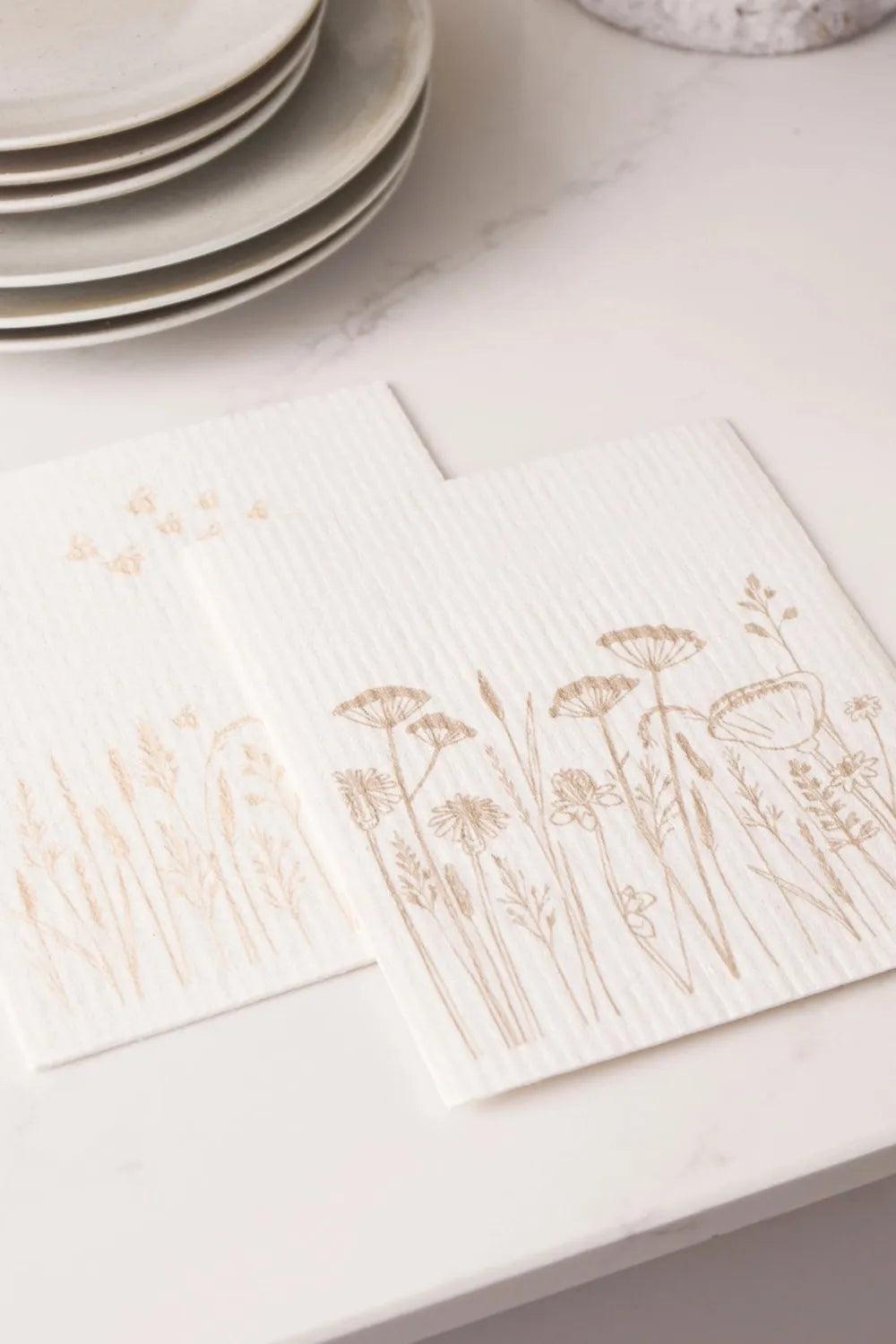 Compostable dishcloths with floral designs on a white surface with plates and a mug.