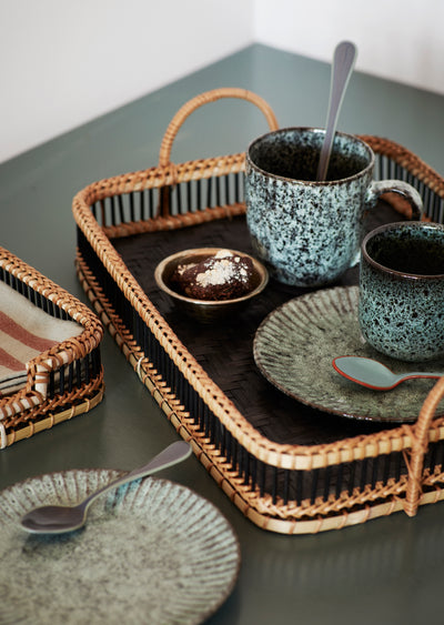 Ceramic mugs and plates on a wicker tray with a spoon on a gray surface.