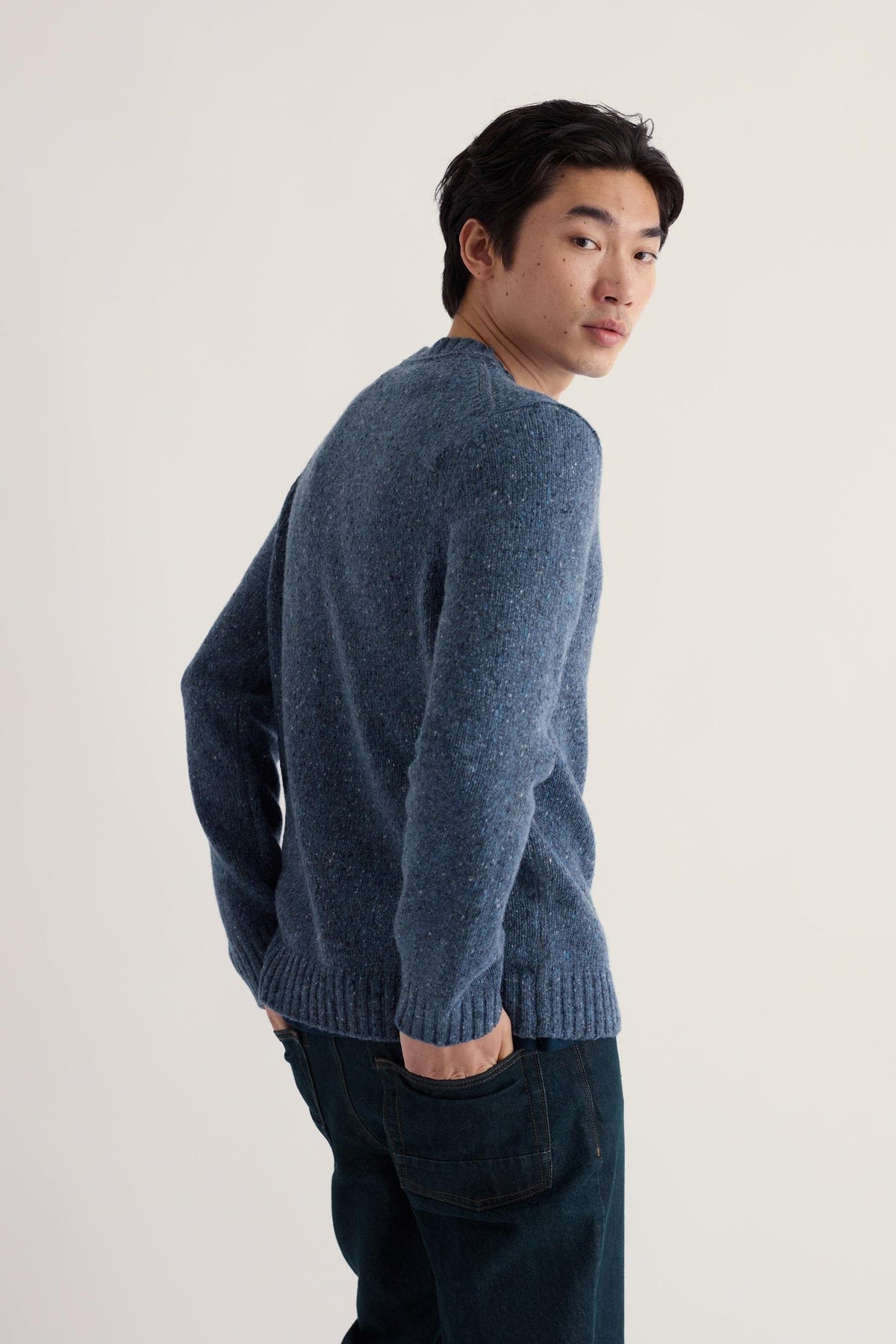 A man looking over his shoulder while wearing a blue Seasalt merino wool jumper against a plain background.