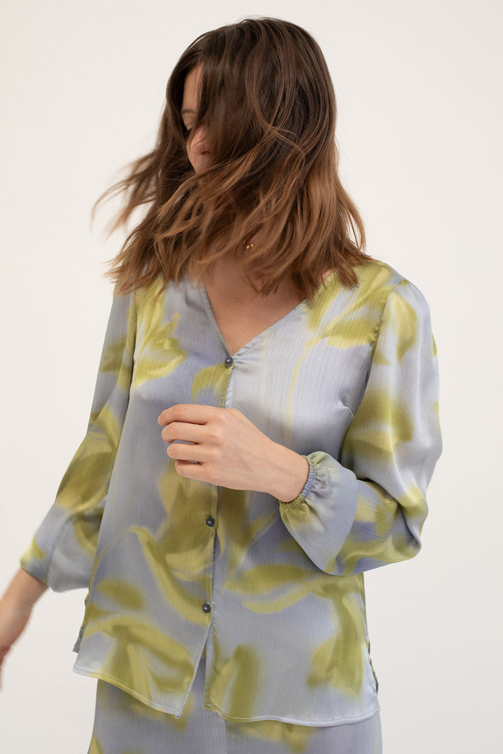Model wearing blouse featuring a satin finish with a subtly textured print. It has a V-neckline, front button closure, and puffed sleeves.