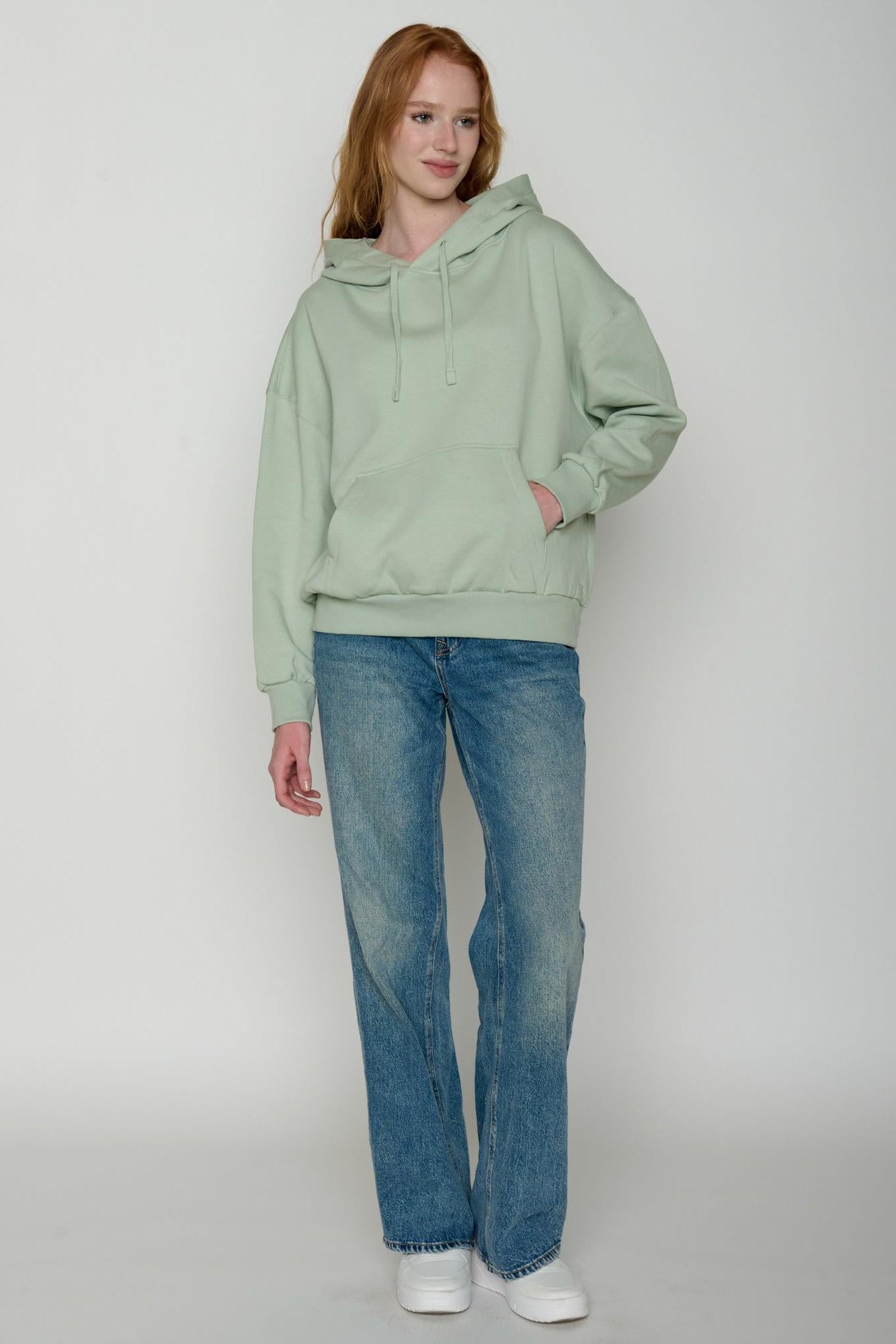 Person wearing a light green 100% organic cotton hoodie and blue jeans on a white background