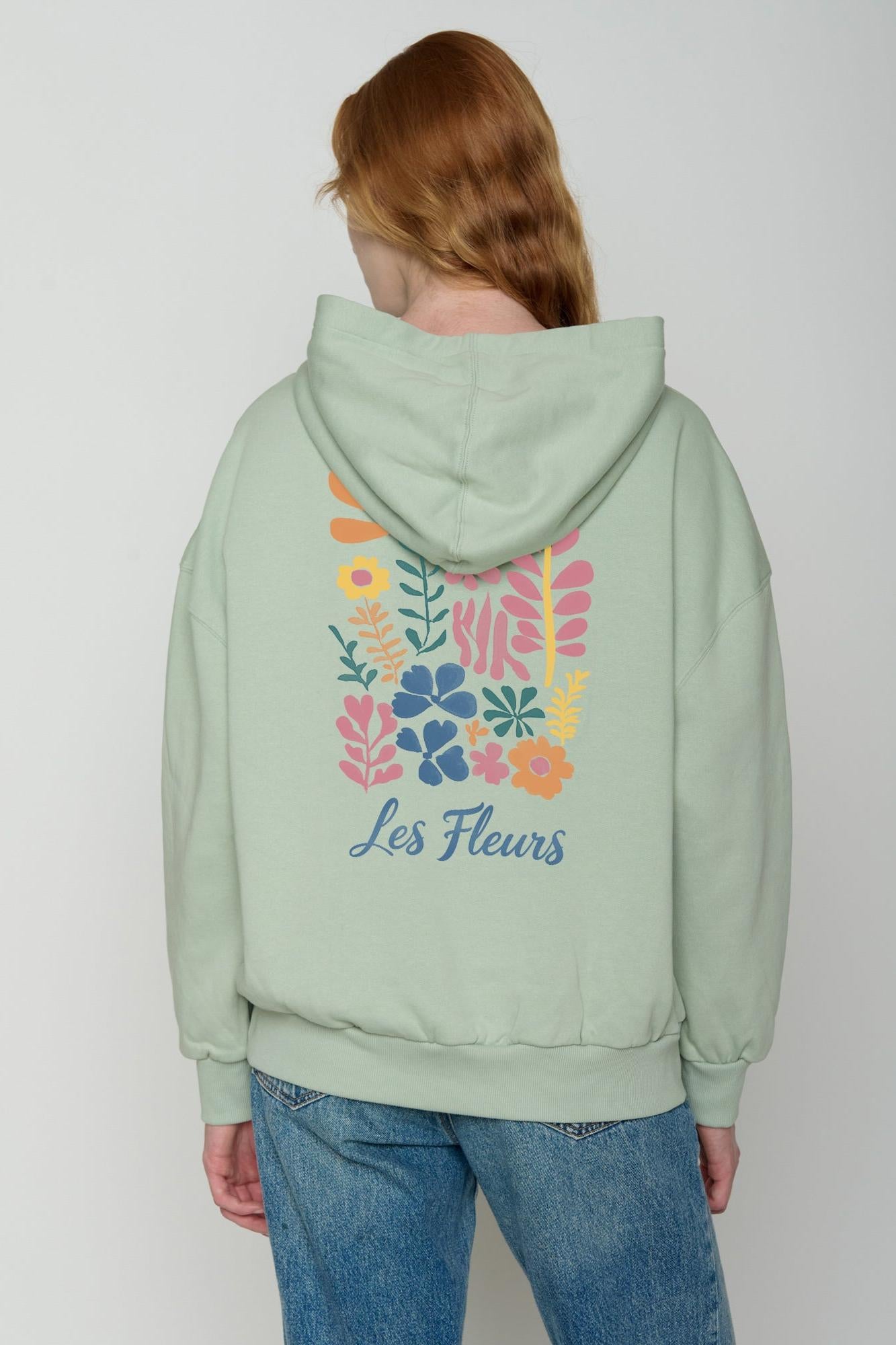 Person wearing a light green hoodie with floral designs and 'Les Fleurs' text on the back, standing against a plain background.