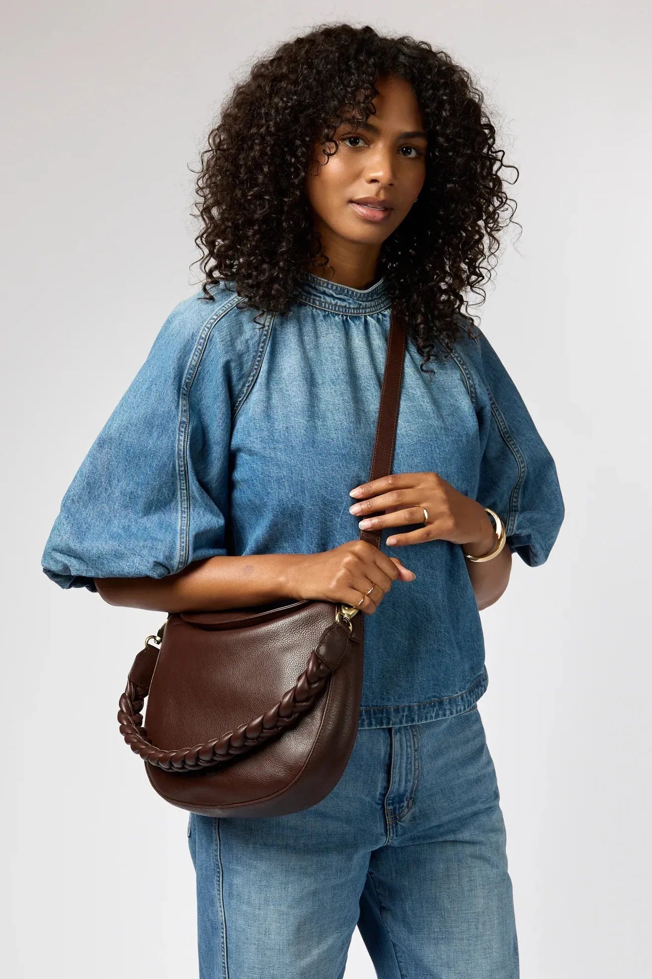 Woman wearing a denim outfit holding a brown leather bag against a plain background